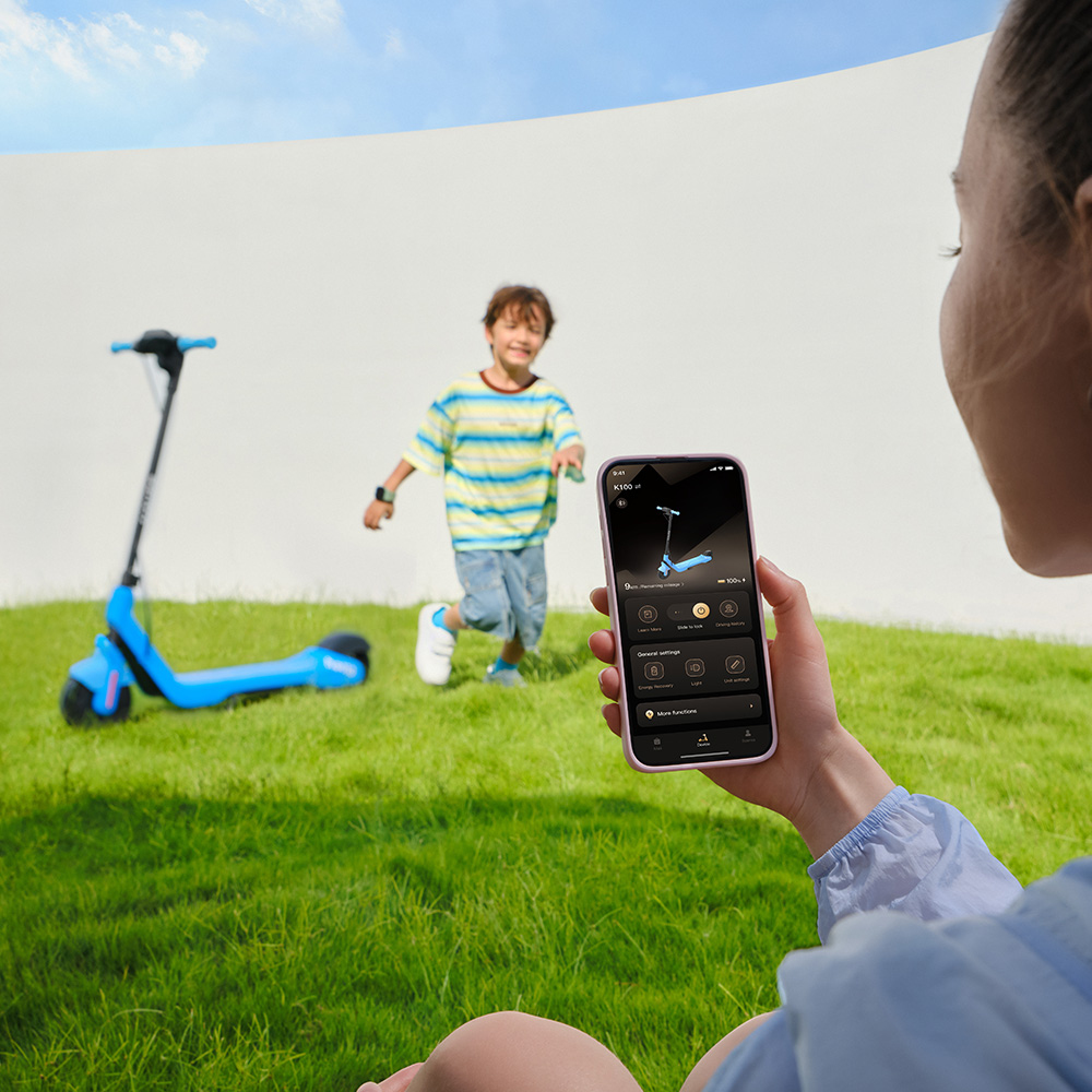 Eine Frau hält ein Smartphone mit einer Roller-App, während im Hintergrund ein Junge neben einem blauen Roller im Gras läuft.