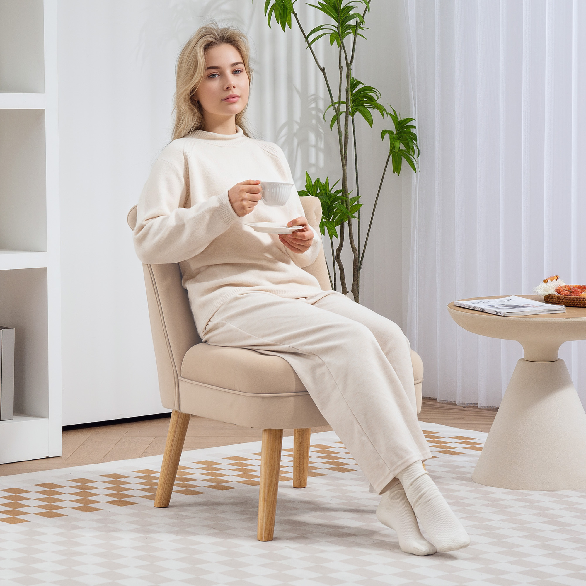 Una donna bionda in abito crema siede su una poltrona, tenendo una tazza in una stanza luminosa.