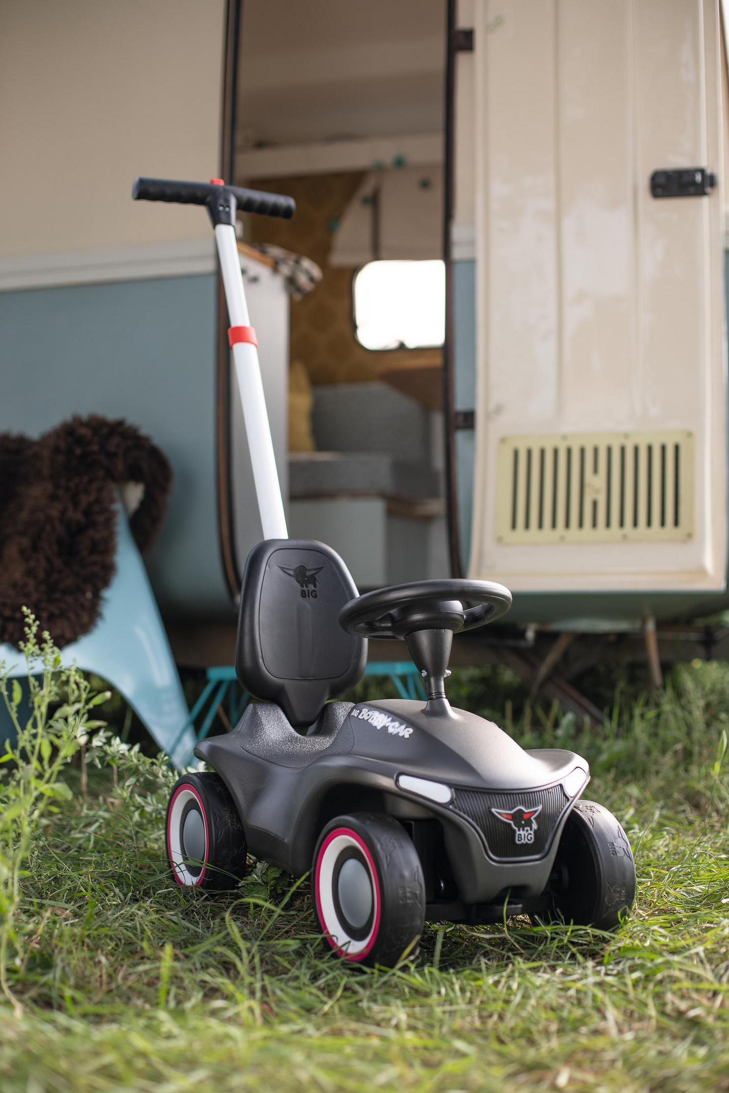 Ein schwarzes Bobby-Car mit Schubstange steht auf einer grünen Wiese vor einem Wohnwagen.