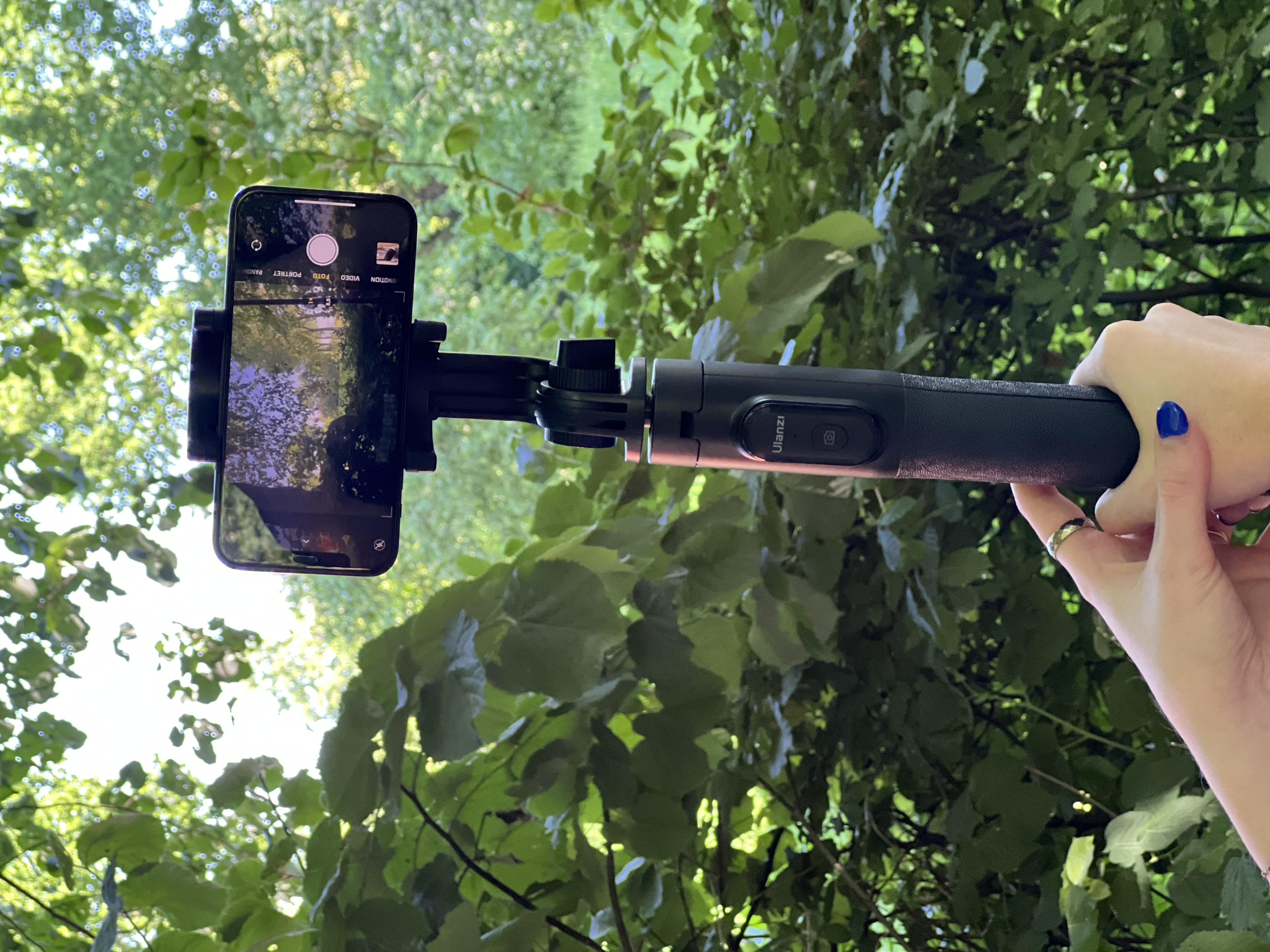 A hand holds a black selfie stick with a smartphone, capturing a view of green trees.