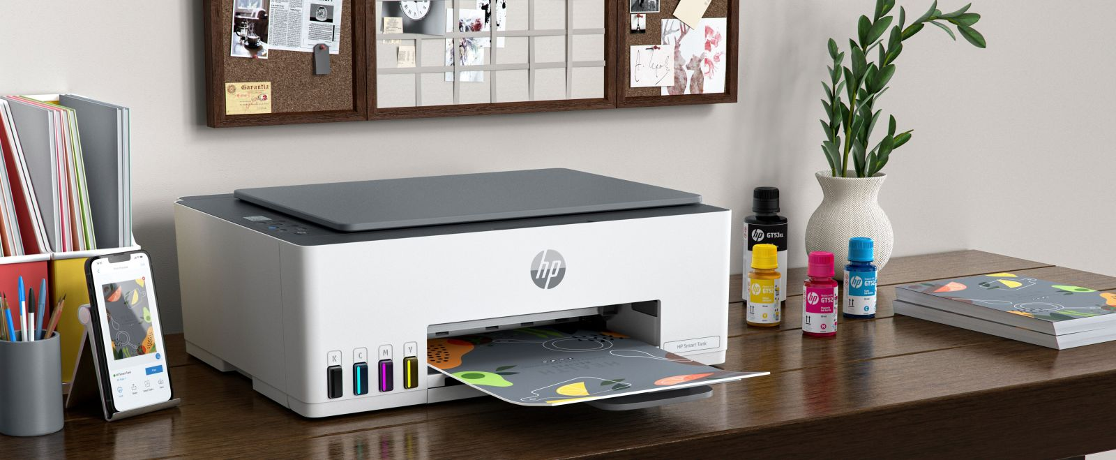 Una stampante HP bianca su una scrivania in legno con flaconi di inchiostro e uno smartphone.