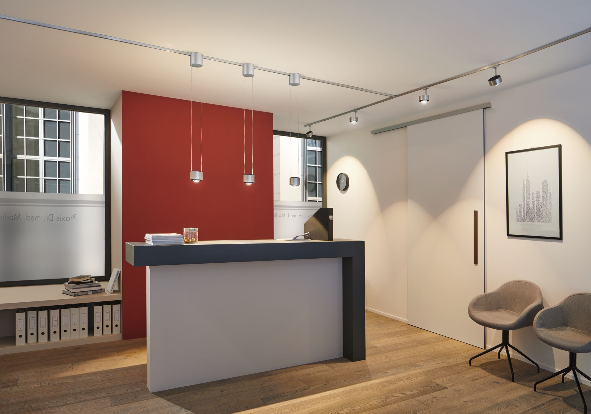 Moderne Rezeption mit roter Wand, grauer Theke, hängenden Lampen, Holzboden und zwei grauen Stühlen.