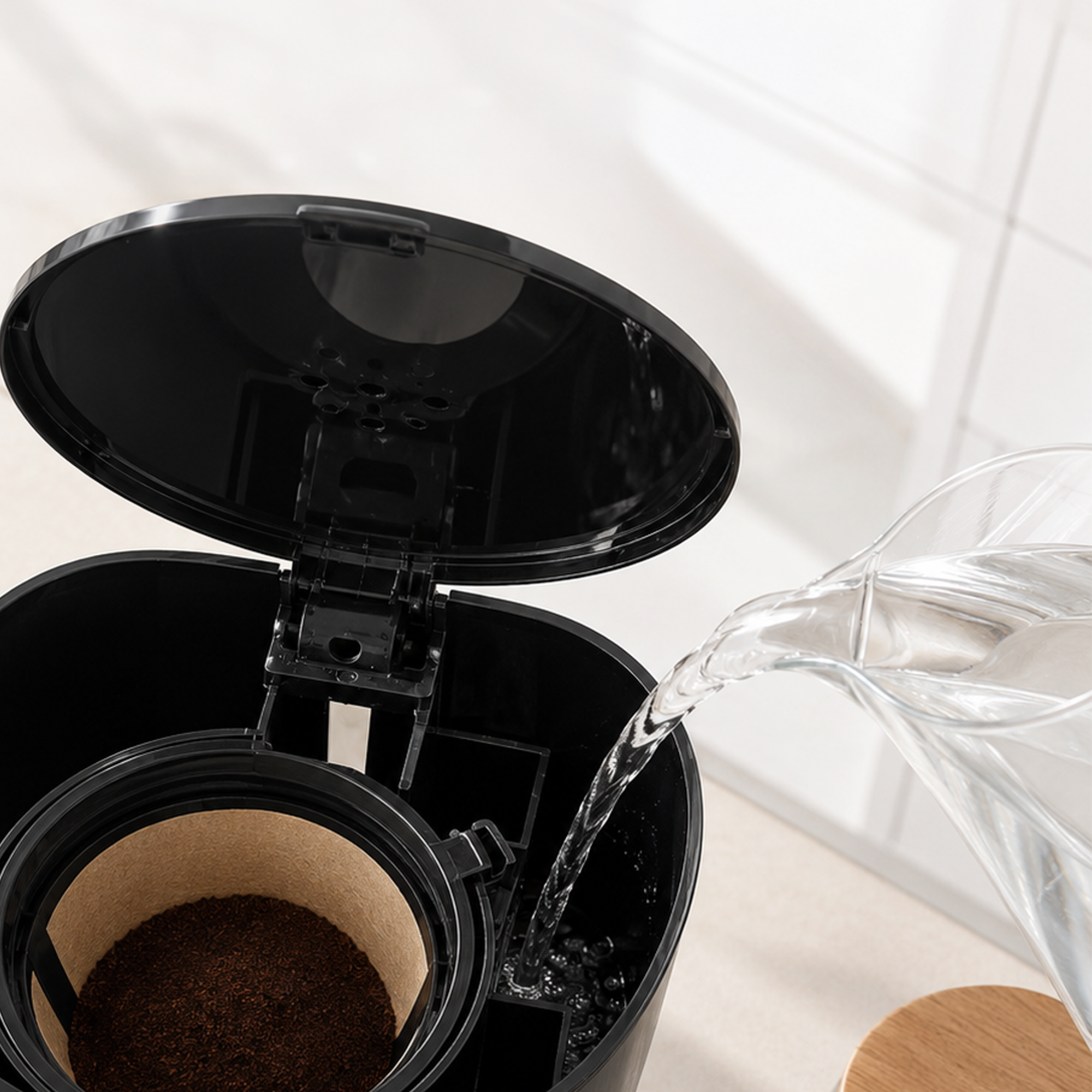 Water is poured from a glass pitcher into a black coffee maker with a paper filter and ground coffee inside.