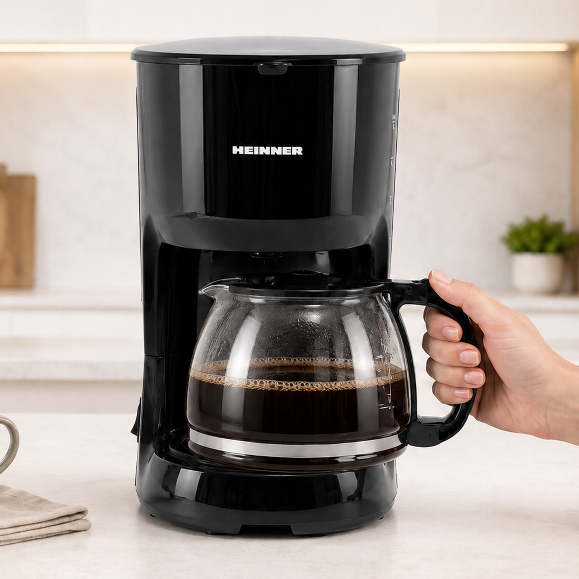 A hand holds a filled glass pot on a black Heinner drip coffee maker on a kitchen counter.
