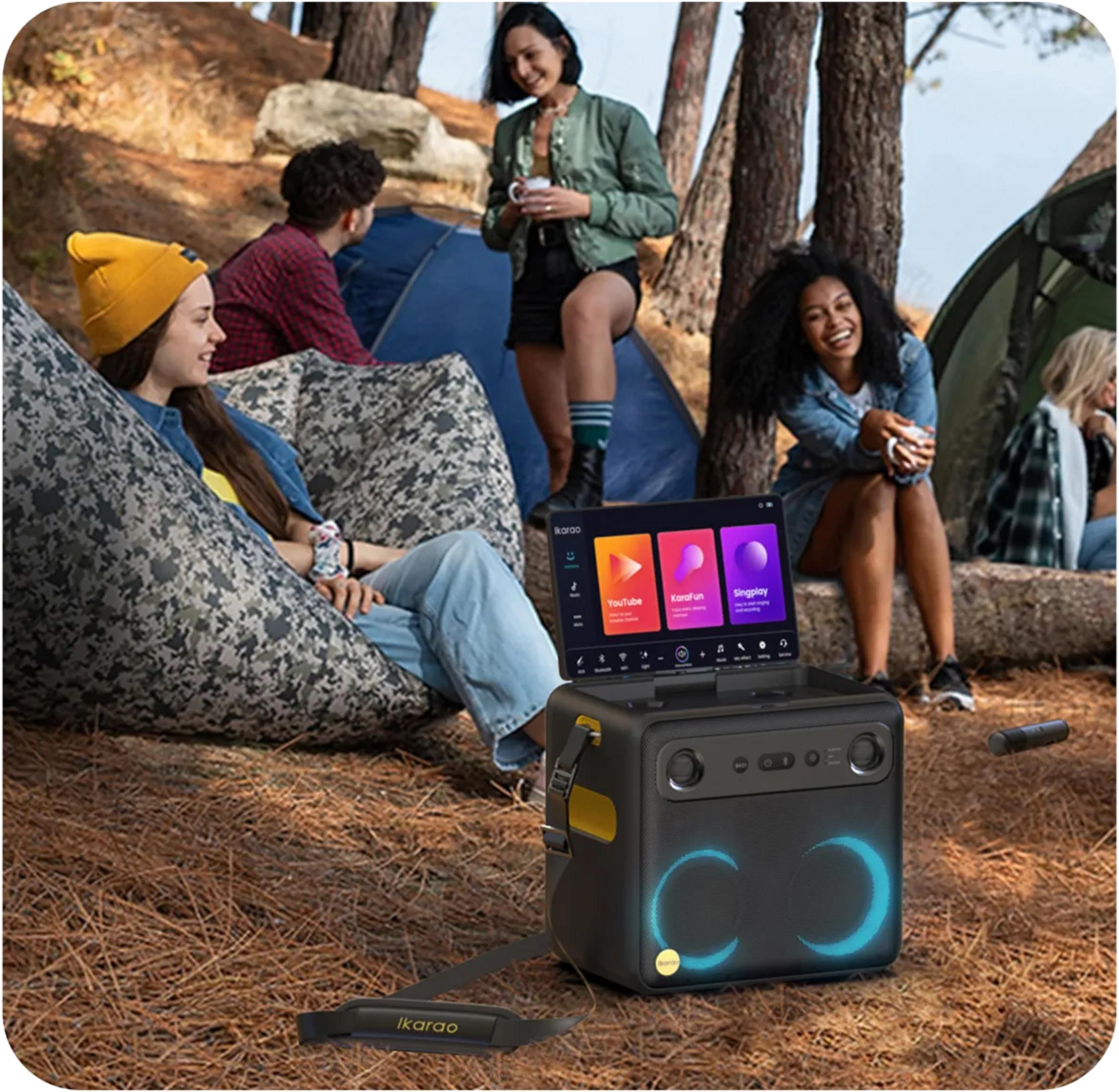 Eine tragbare Karaoke-Maschine im Wald, mit einer Gruppe von Freunden, die im Hintergrund campen und entspannen.
