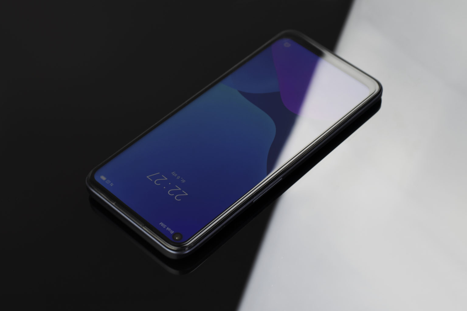 Smartfon leży na czarno-białej powierzchni, wyświetlając niebieską tapetę.