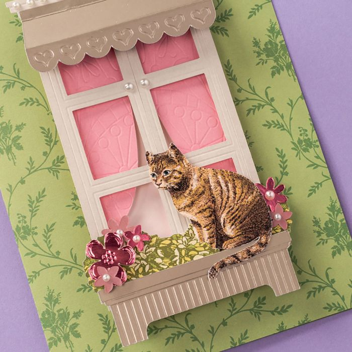 Eine handgemachte Papierkarte zeigt eine Katze auf einem Fensterbrett mit rosafarbenem Vorhang und Blumen.
