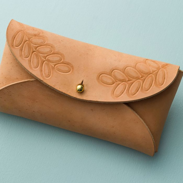 Ein hellbraunes Lederetui mit Blattmustern und einem goldenen Knopfverschluss auf blauer Oberfläche.