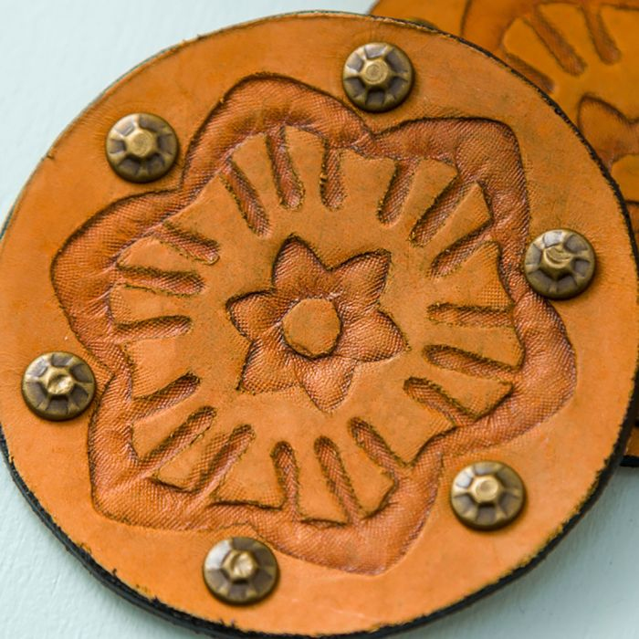 Runder brauner Lederuntersetzer mit geprägtem Blumenmuster und Metallnieten am Rand auf hellblauer Oberfläche.