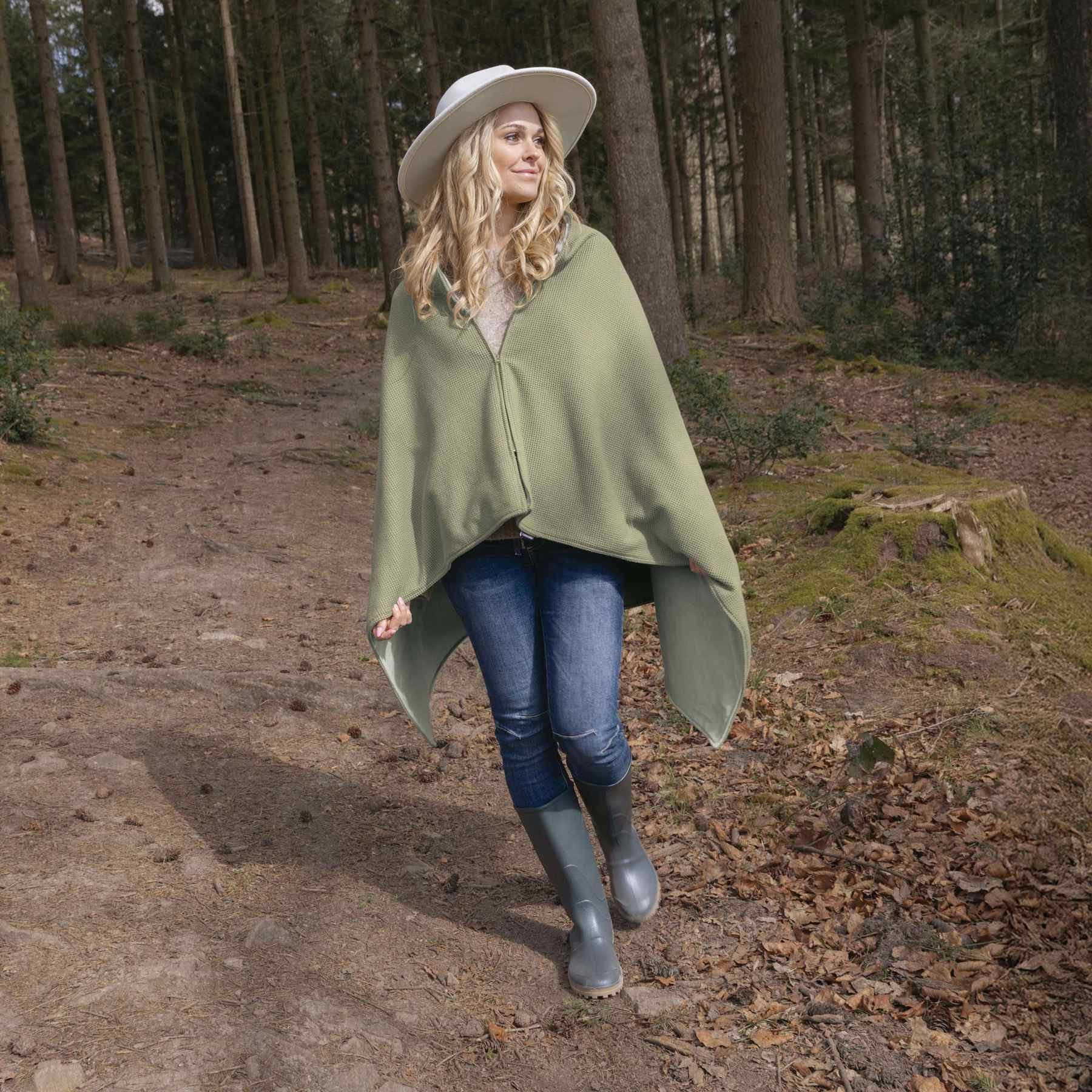 Blonde woman wearing a hat, green cape, jeans, and rubber boots walking on a forest path.