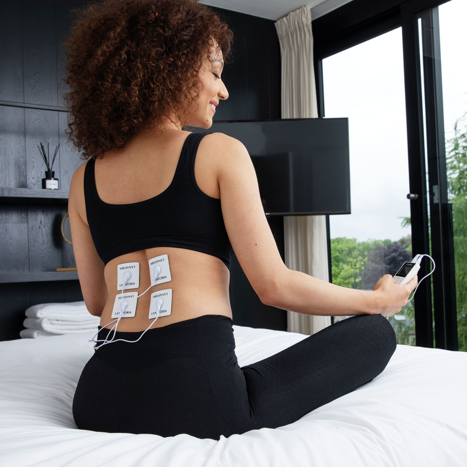 Woman sits on a bed with TENS unit pads on her lower back, holding a remote.