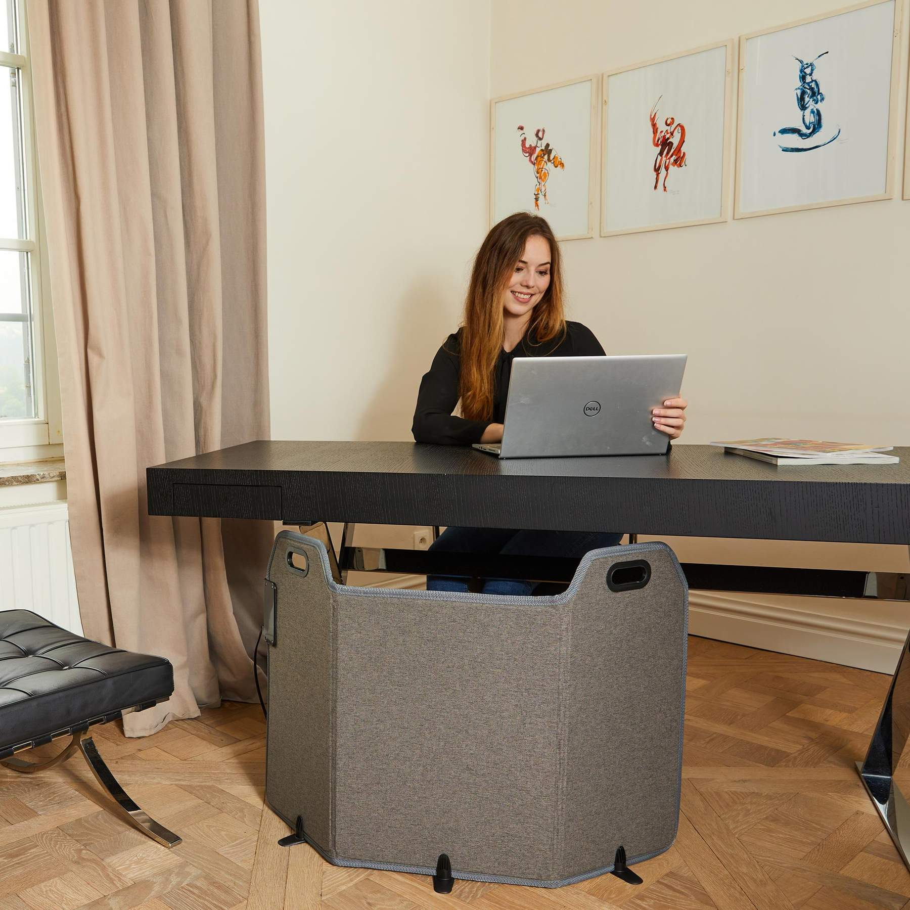 Eine Frau arbeitet an einem Laptop an einem dunklen Holztisch mit einem grauen Stoff-Heizpanel darunter.