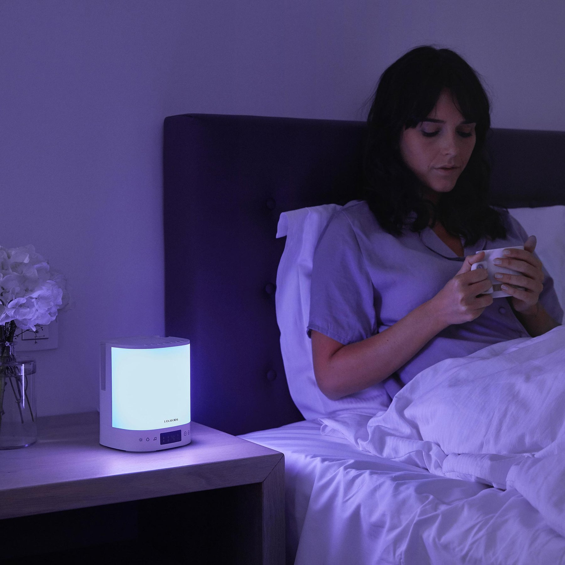 Eine Frau im Bett hält eine Tasse, neben ihr auf dem Nachttisch steht eine hell leuchtende Lampe.