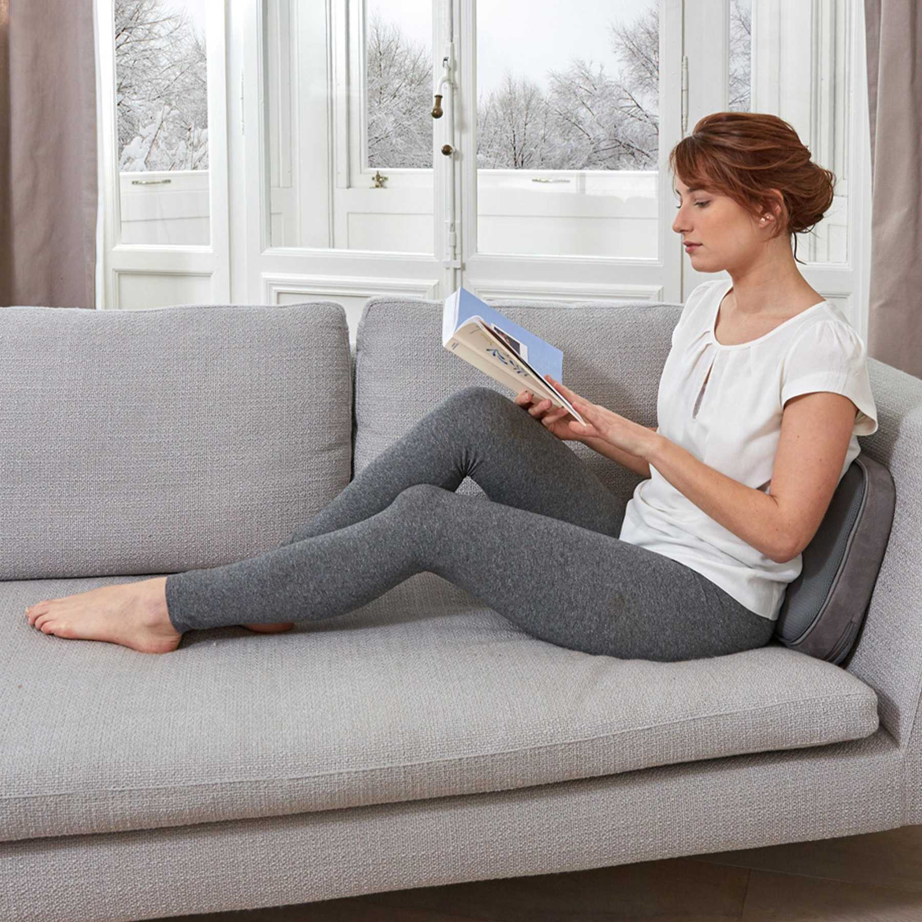 Eine Frau liest ein Buch auf einem grauen Sofa vor einem verschneiten Fenster.