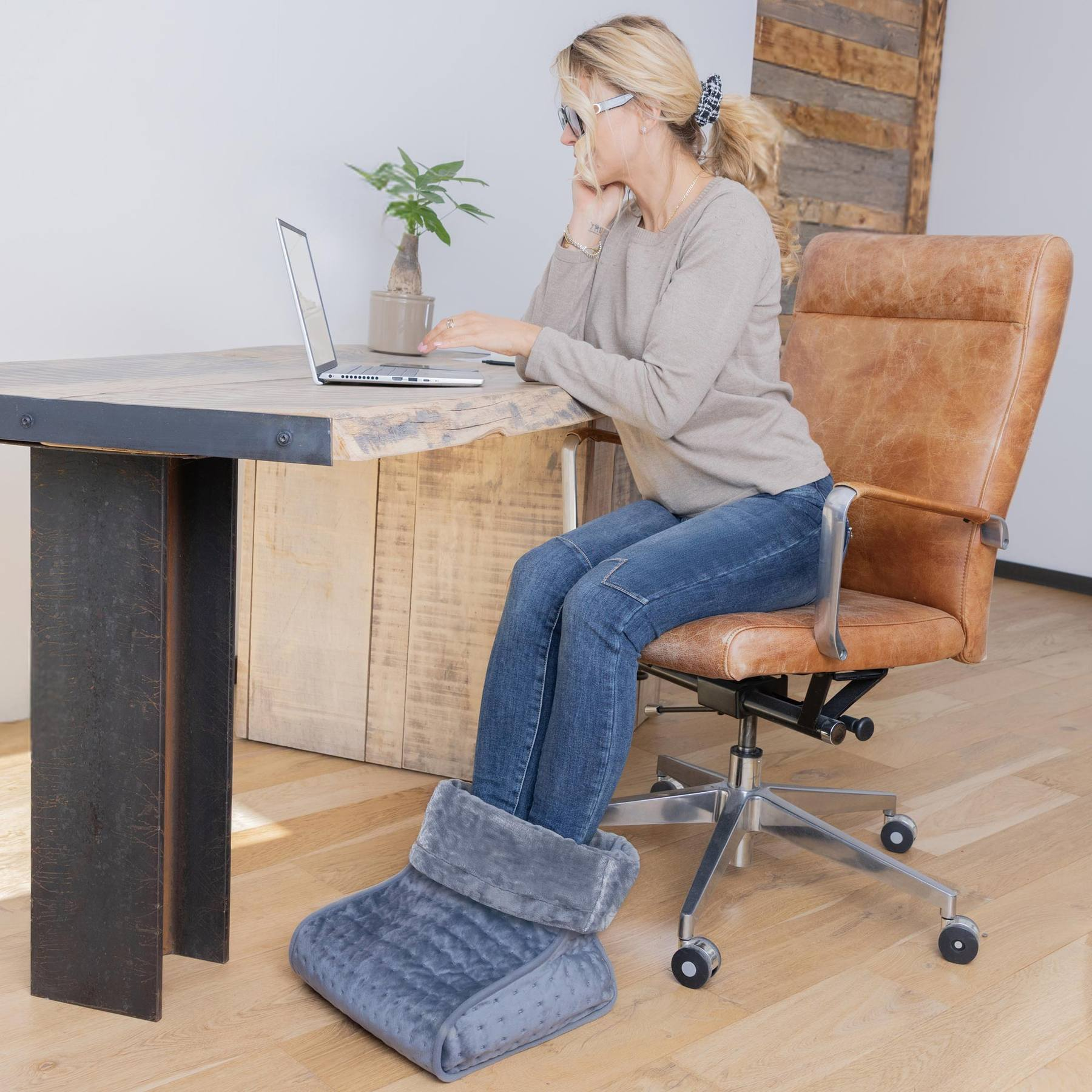 A woman works on a laptop at a wooden desk while her feet are inside a gray foot warmer.