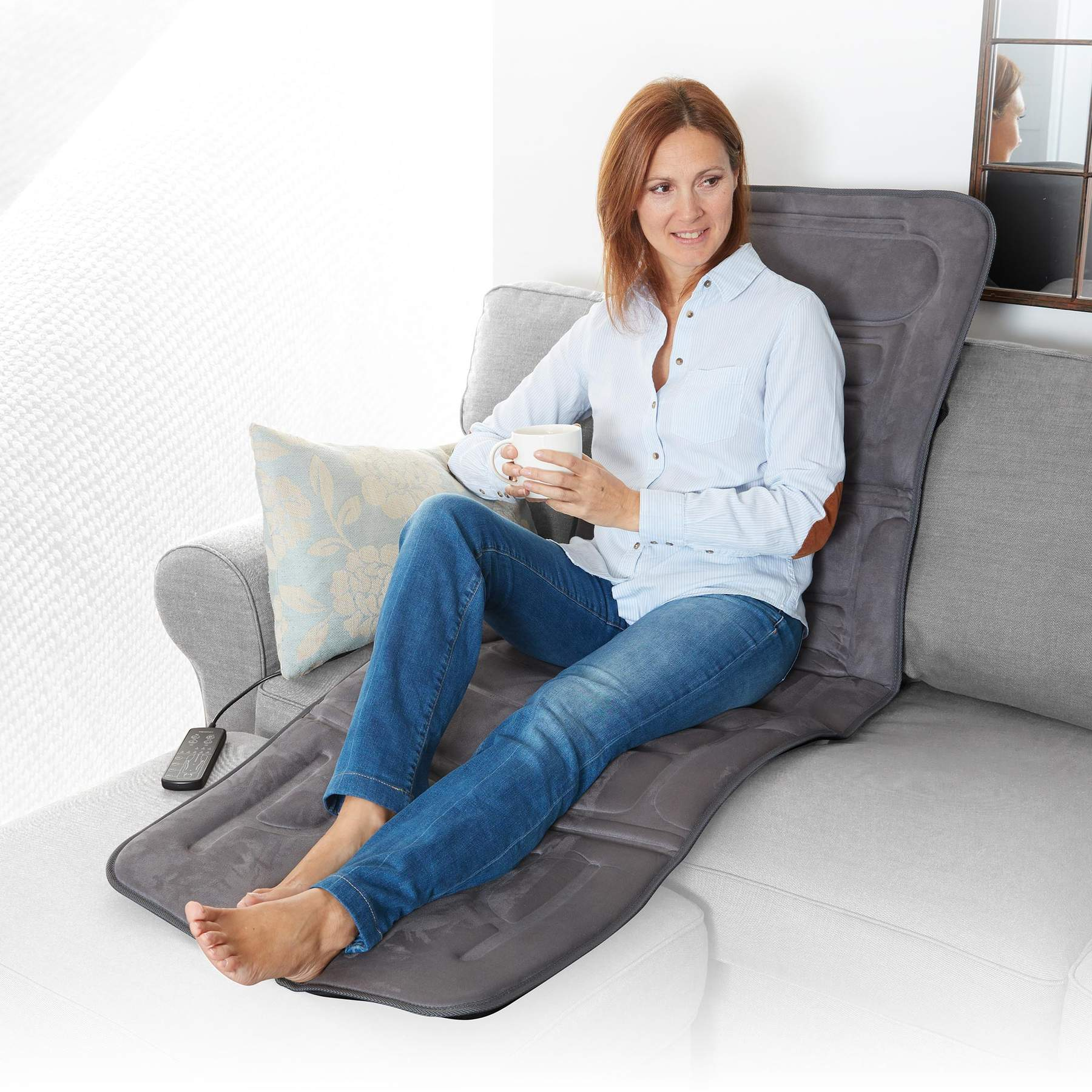 Eine Frau sitzt auf einem Sofa mit einer grauen Massageauflage, hält eine Tasse und schaut nach rechts.