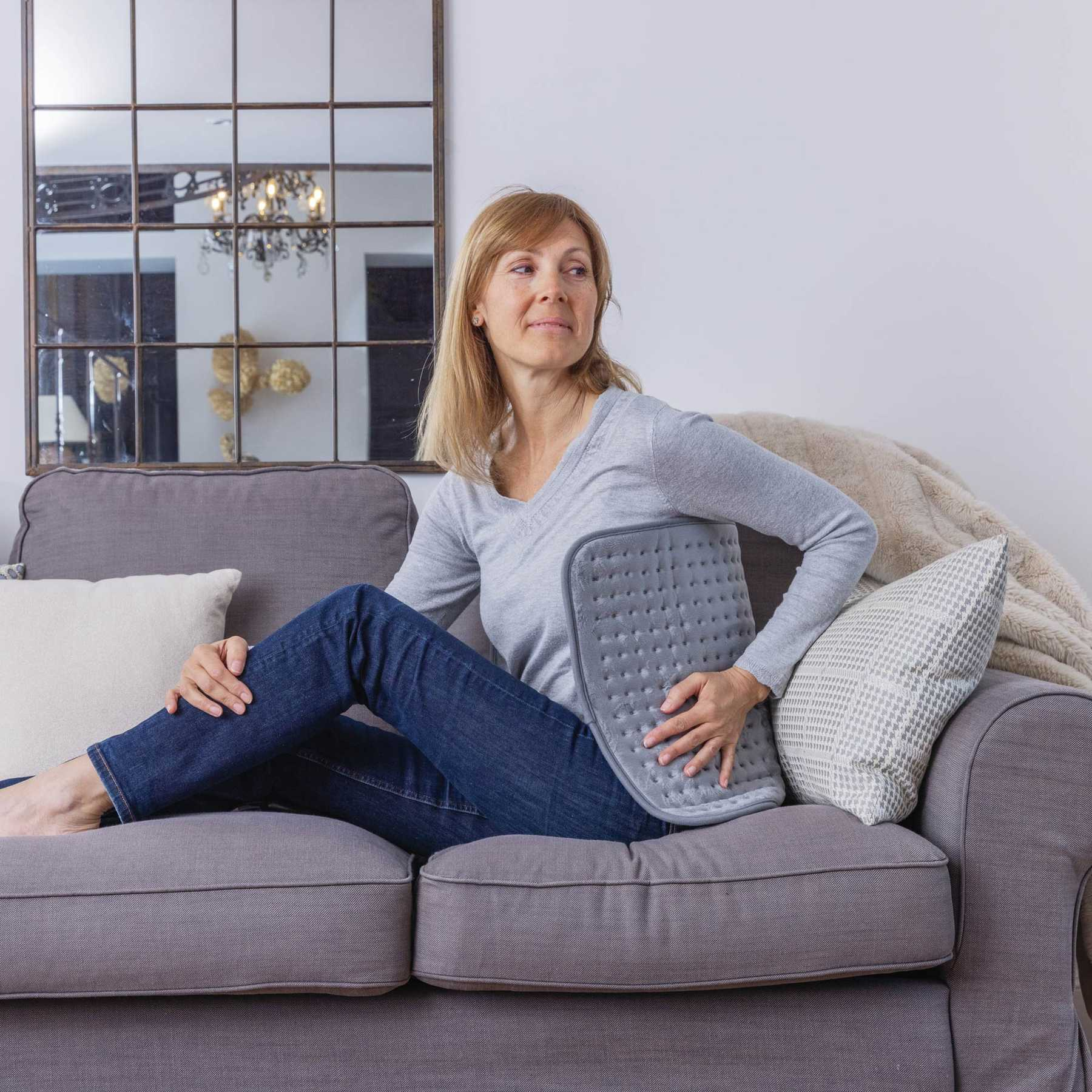 Eine blonde Frau sitzt auf einem grauen Sofa und hält ein graues Wärmekissen gegen ihren Rücken.