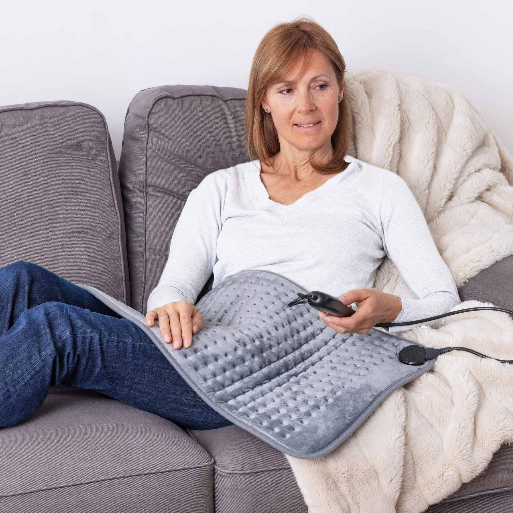 Eine Frau sitzt auf einem grauen Sofa und hält ein graues elektrisches Heizkissen auf ihrem Schoß mit einer Fernbedienung.