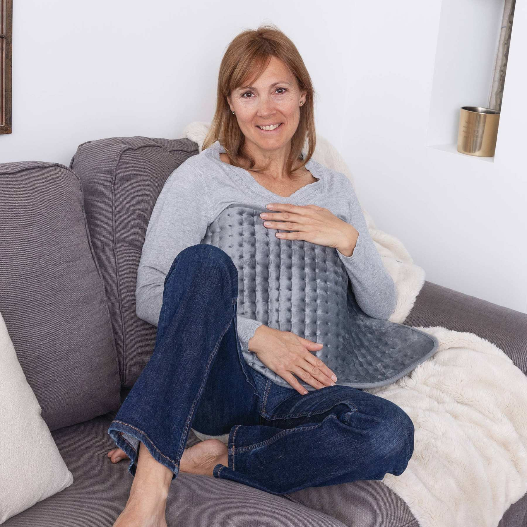 Eine Frau sitzt auf einem grauen Sofa und hält ein graues Heizkissen auf ihrem Bauch.