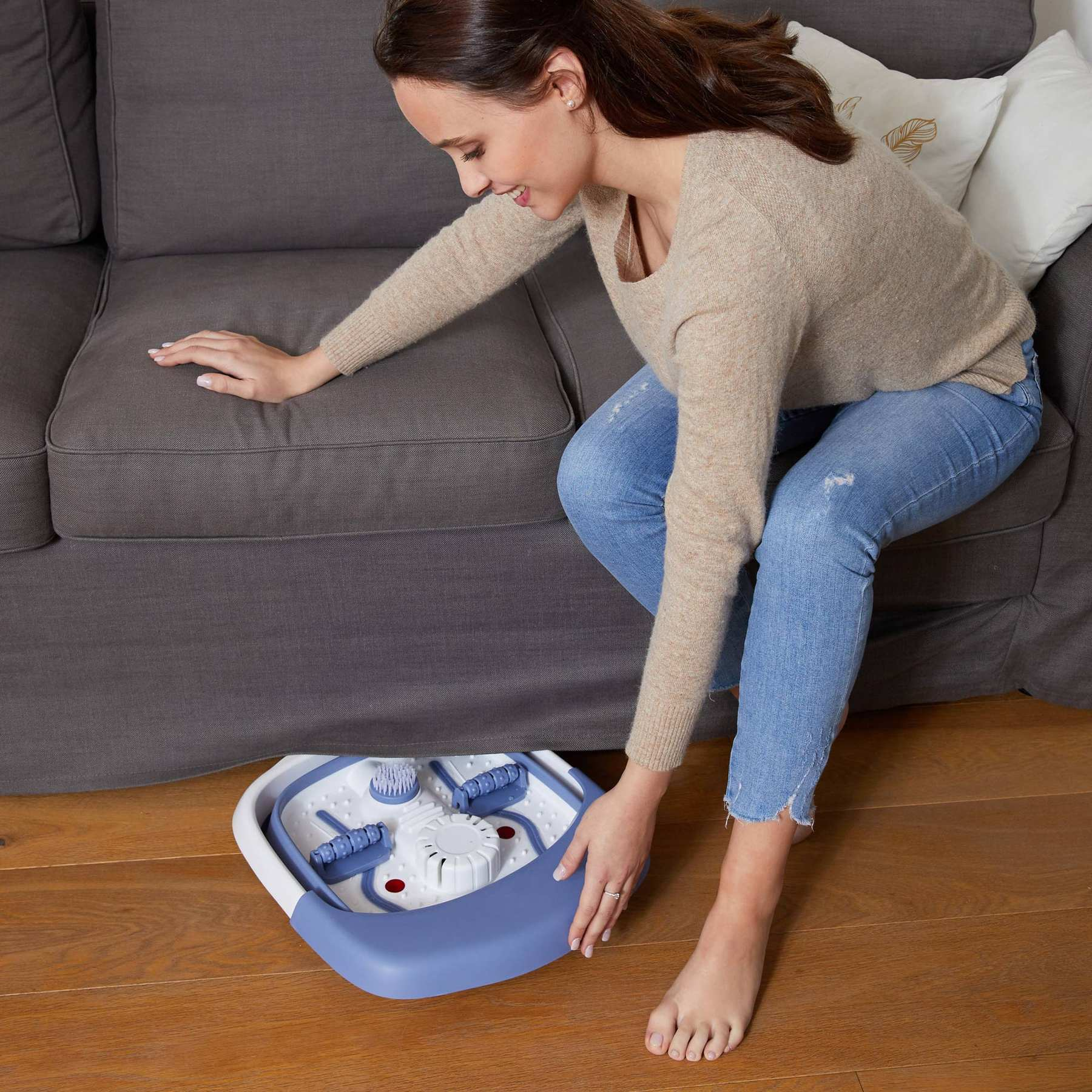 A woman in a beige sweater places a blue and white foot spa on the wooden floor in front of a grey sofa.