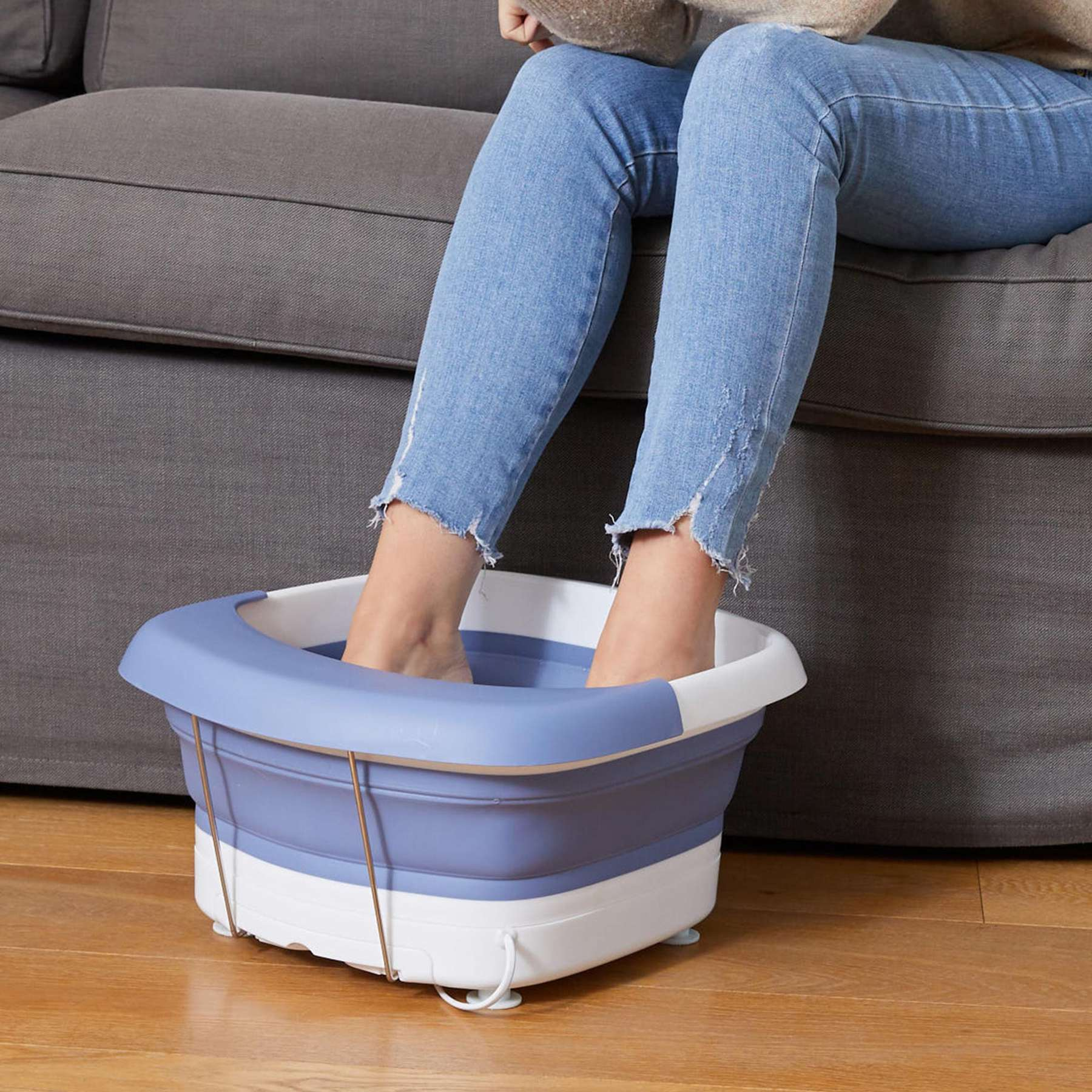 Person sits on a grey sofa, soaking feet in a blue and white foot bath placed on a wooden floor.