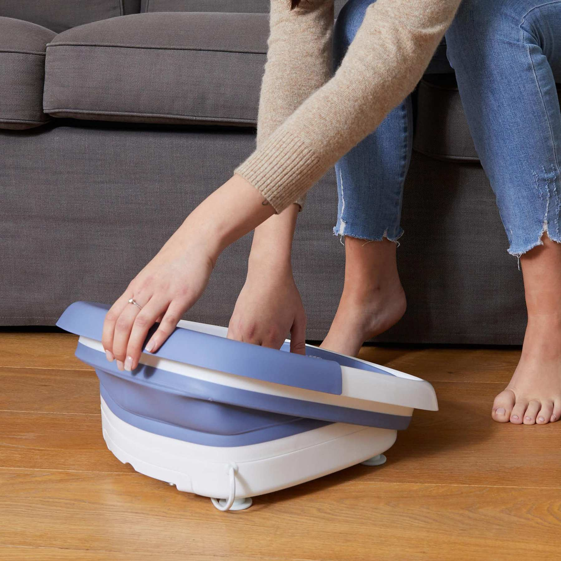 A person sits on a grey sofa, setting up a blue and white collapsible foot basin on a wooden floor.