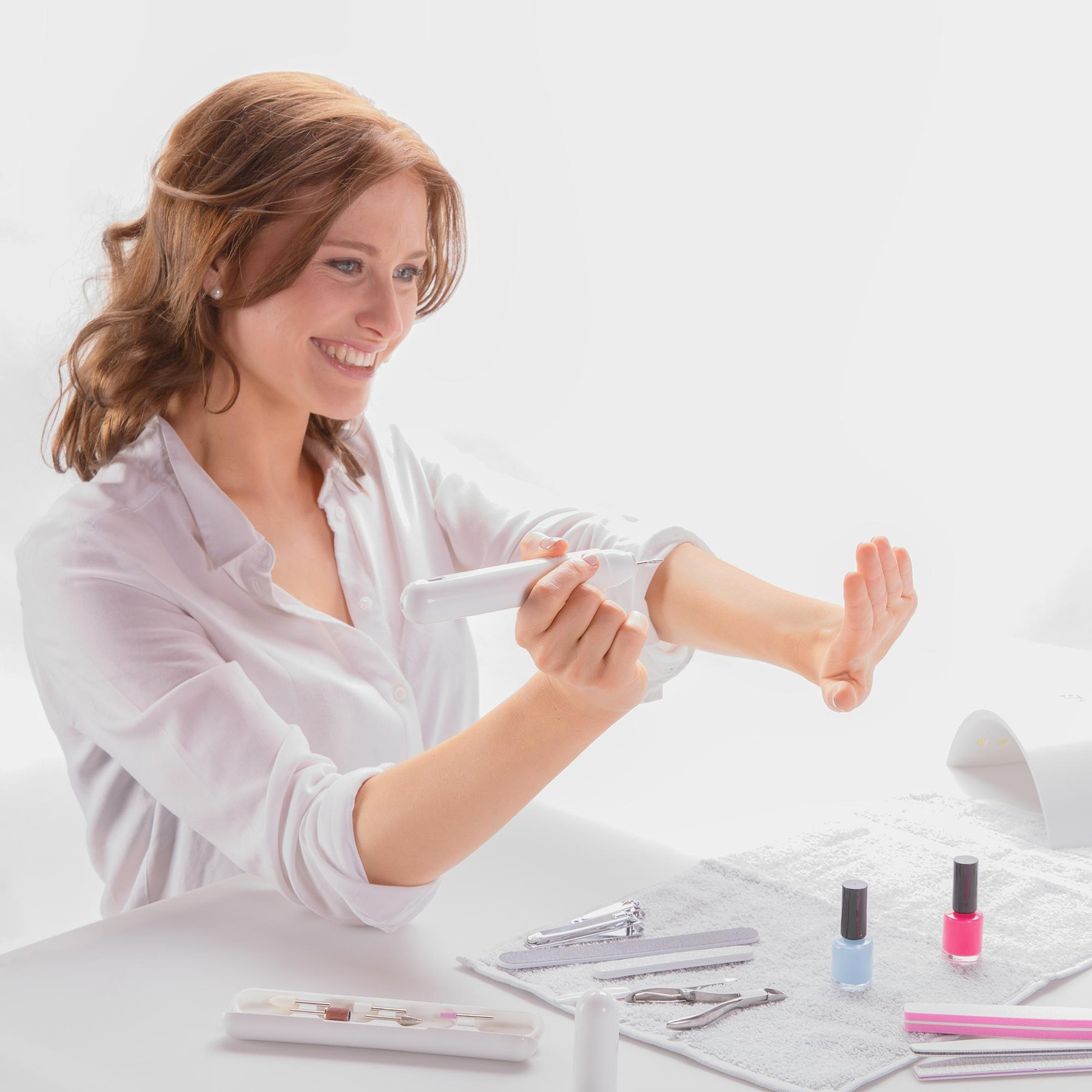 Eine lächelnde Frau benutzt ein elektrisches Nagelset an ihrem Arm, umgeben von Nagellacken und Feilen.