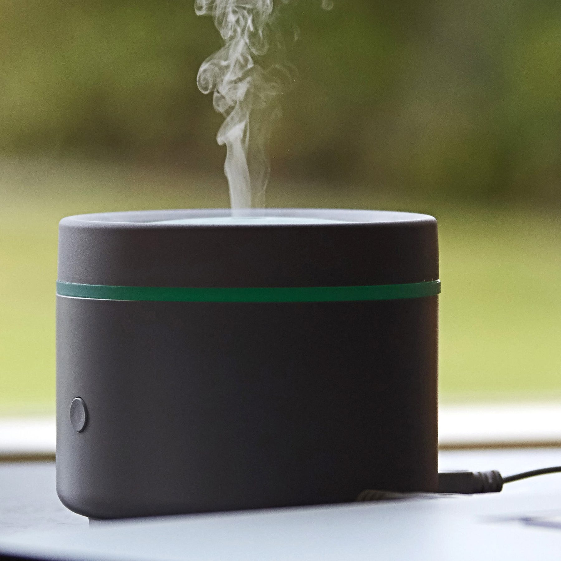 A grey diffuser on a table releases steam, featuring a green ring and a blurred outdoor background.