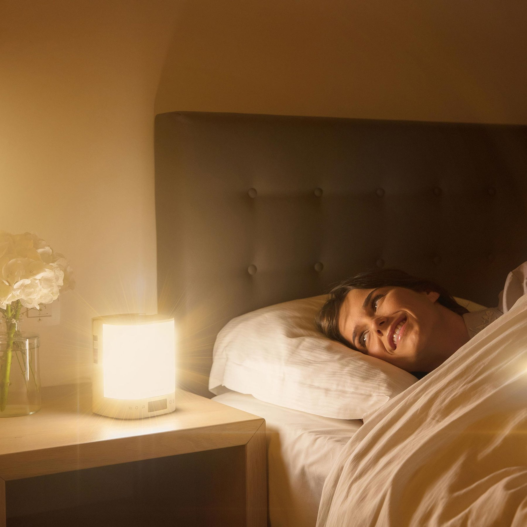 Eine lächelnde Frau liegt im Bett, beleuchtet von einem hellen Nachtlicht auf ihrem Nachttisch.
