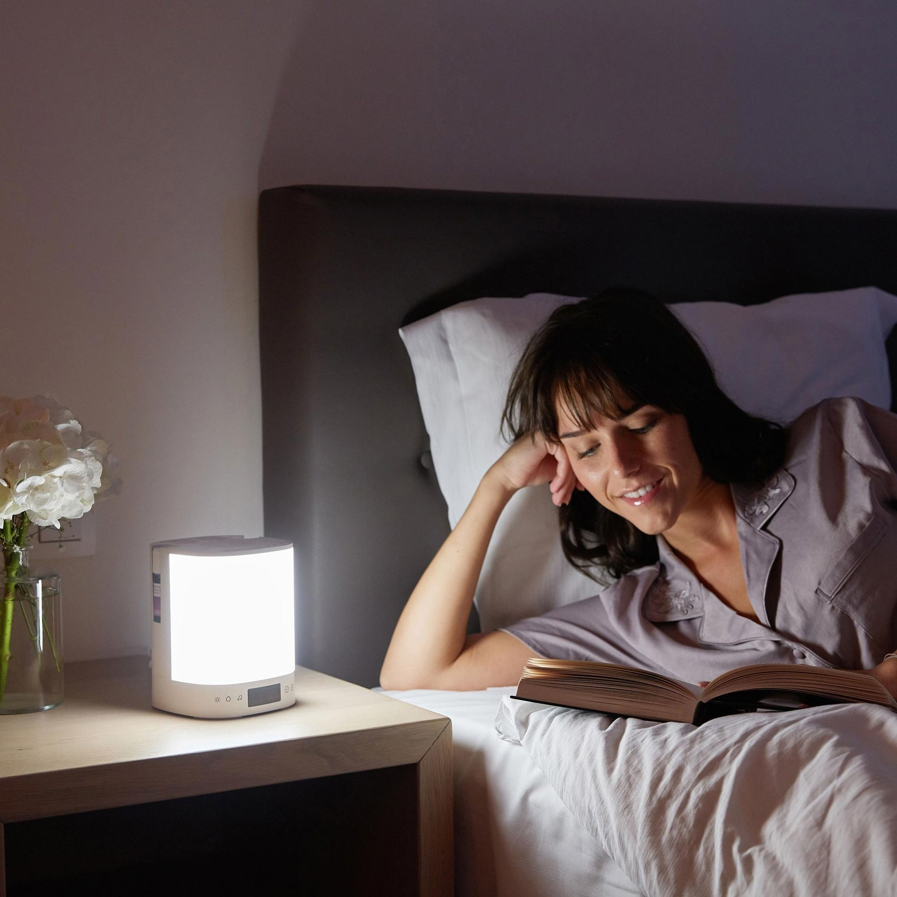 Eine lächelnde Frau liest im Bett neben einer Nachttischlampe und Blumen ein Buch.