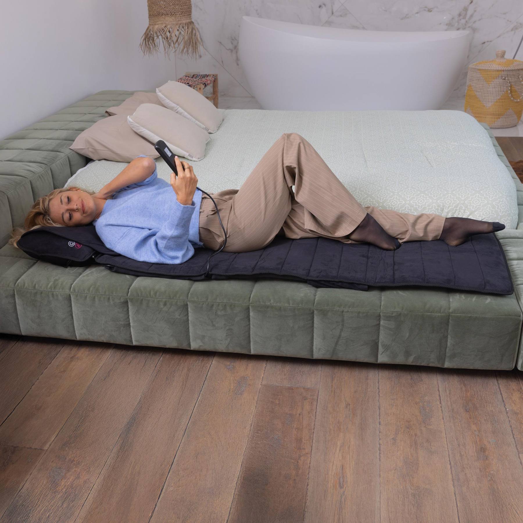 A blonde woman lies on a black massage mat atop a large green bed, holding a remote control.
