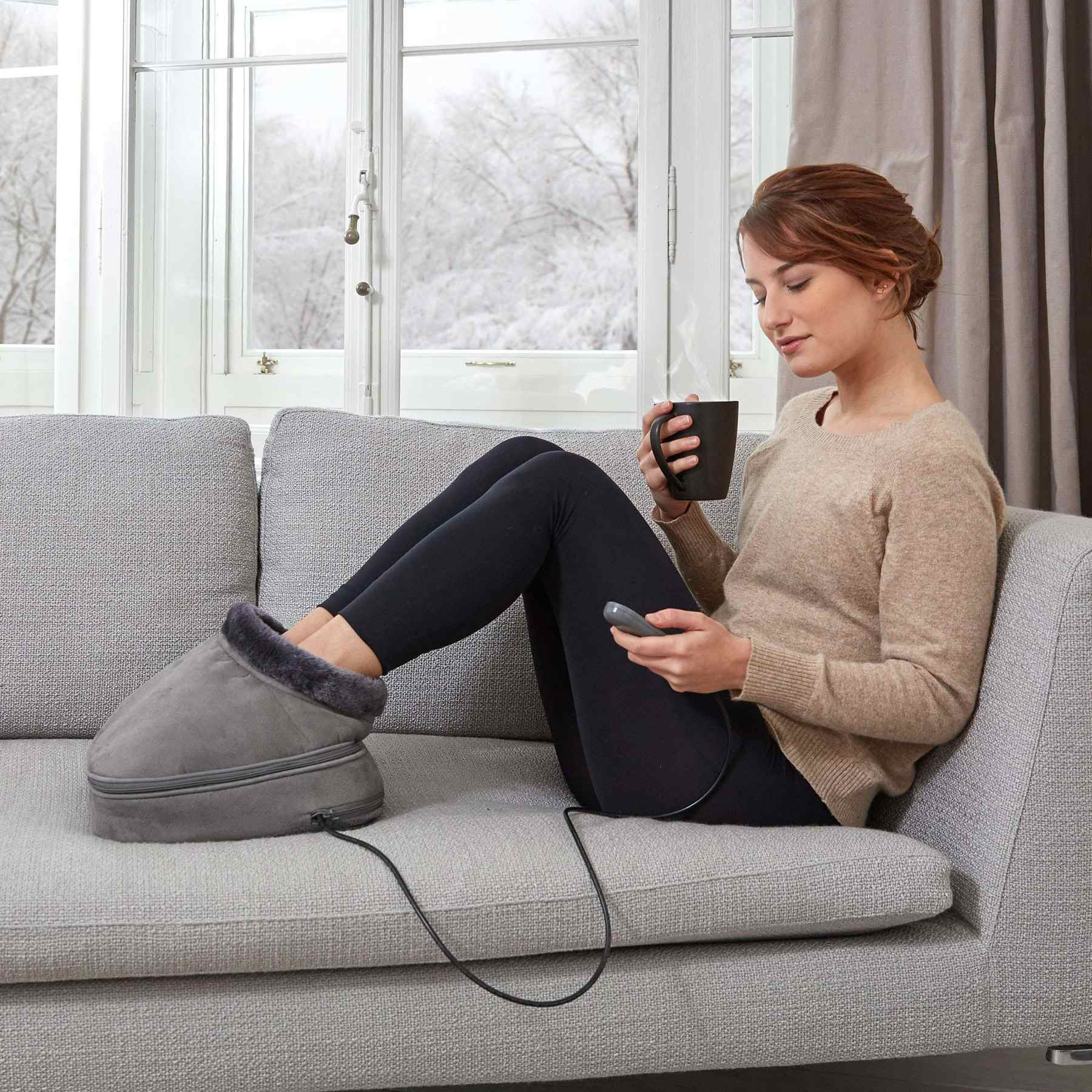 Eine Frau sitzt auf einem Sofa, hält eine Tasse, während ihre Füße in einem elektrischen Fußwärmer stecken.