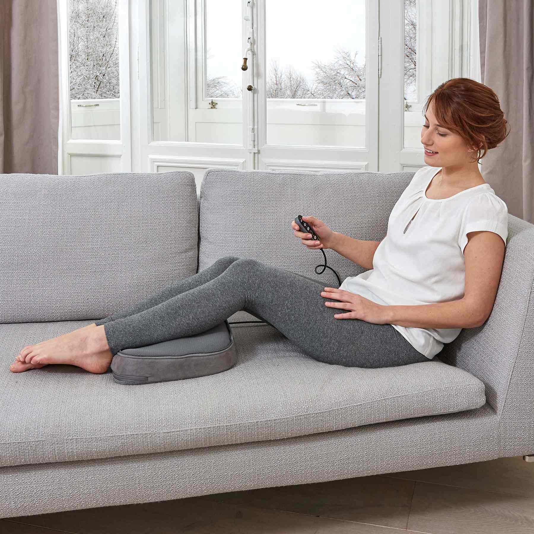 Eine Frau sitzt auf einem Sofa und hält eine Fernbedienung für ein Fußmassagegerät in der Hand.