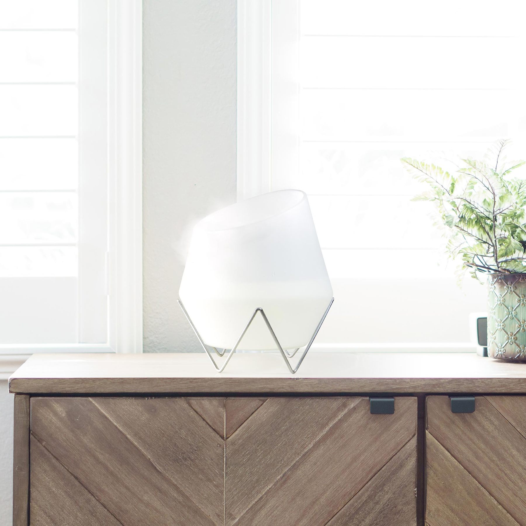 A white diffuser on a metal stand rests on a wooden cabinet, with a potted plant nearby against a bright window.
