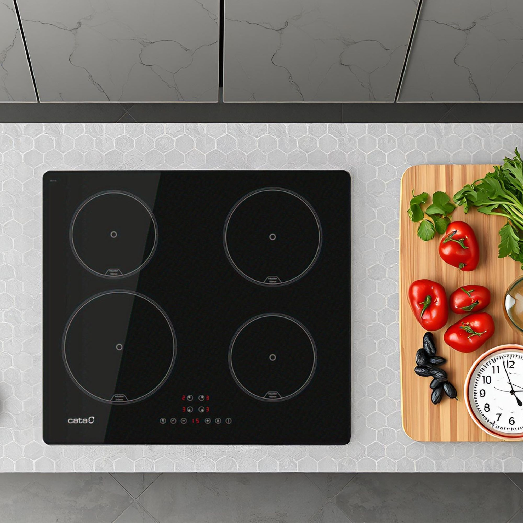 Una placa de inducción negra y una tabla de cortar de madera con tomates, hierbas, aceitunas y un reloj en el mostrador.
