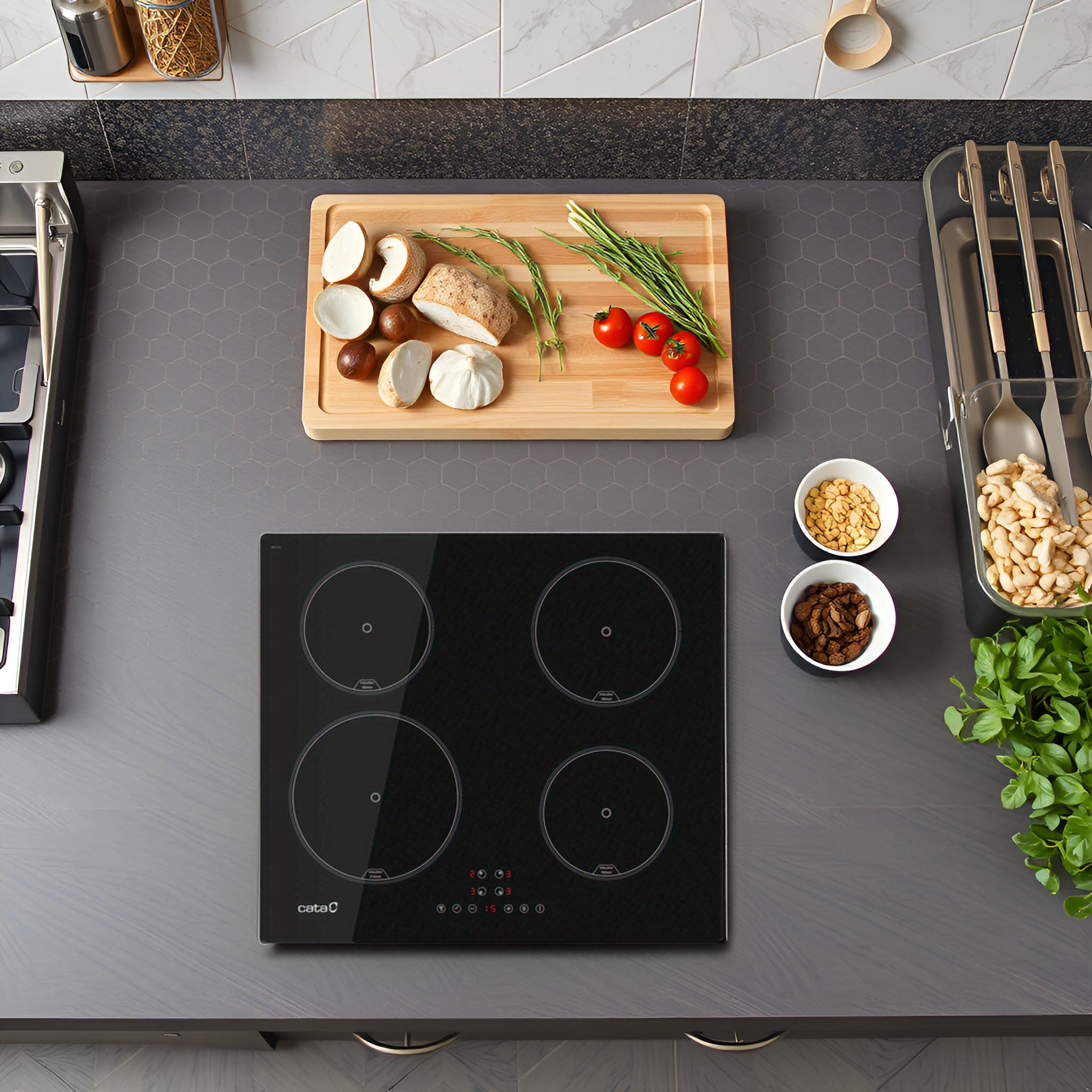 Una encimera de cocina con placa de inducción negra, tabla con verduras y cuencos con frutos secos.