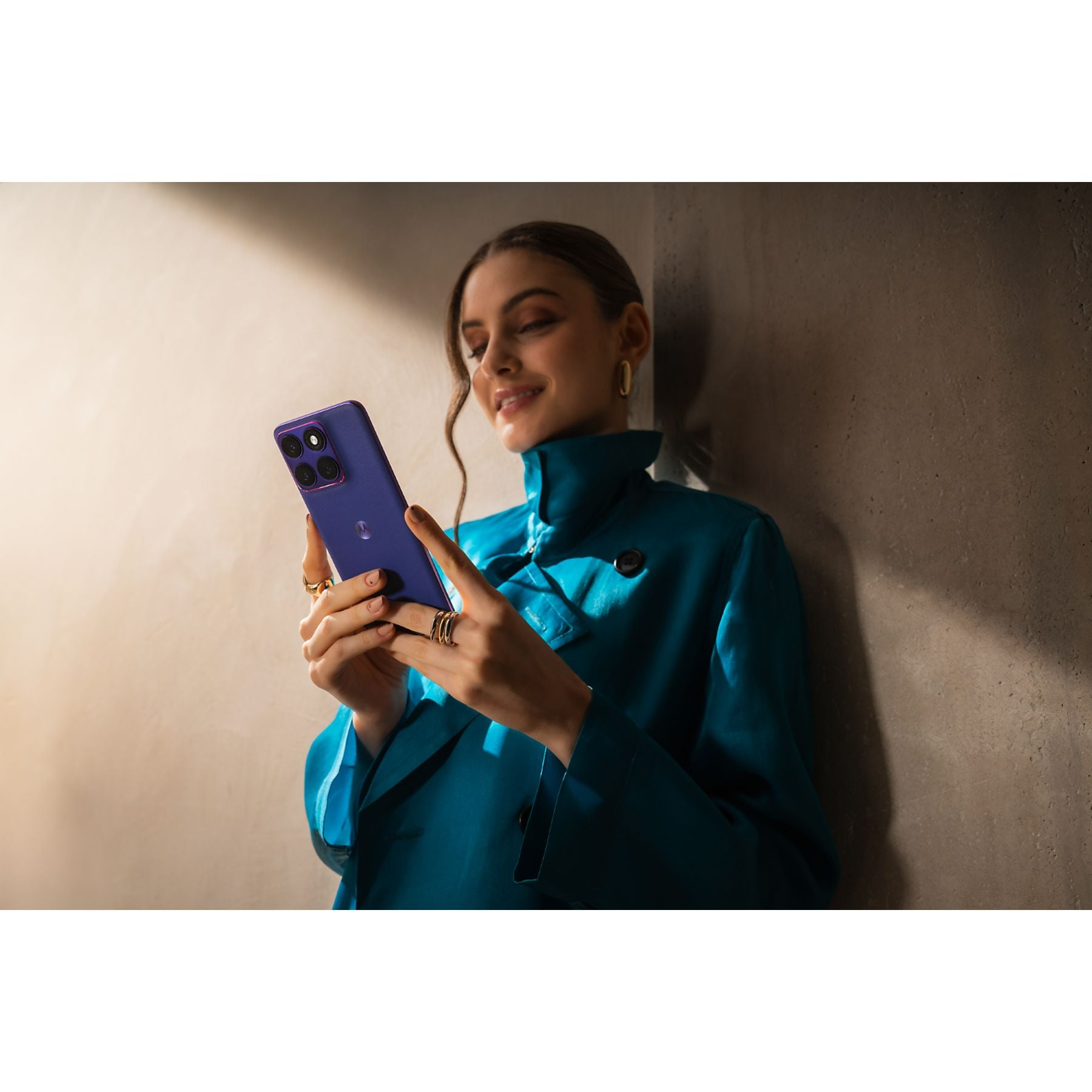 Una mujer con abrigo azul sostiene un teléfono inteligente morado ante una pared.