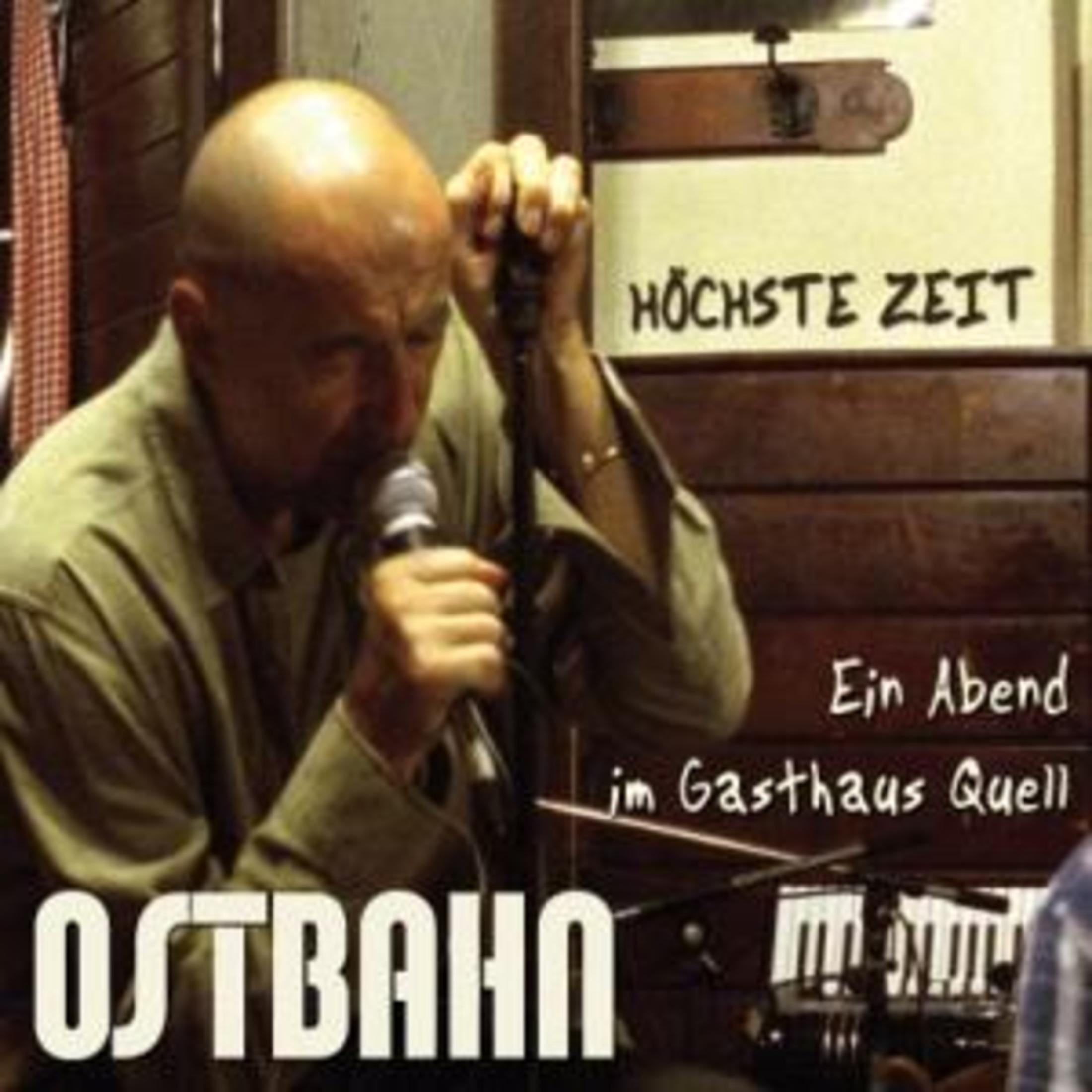 Ein kahlköpfiger Mann singt in ein Mikrofon, im Hintergrund Holzvertäfelung und Text.