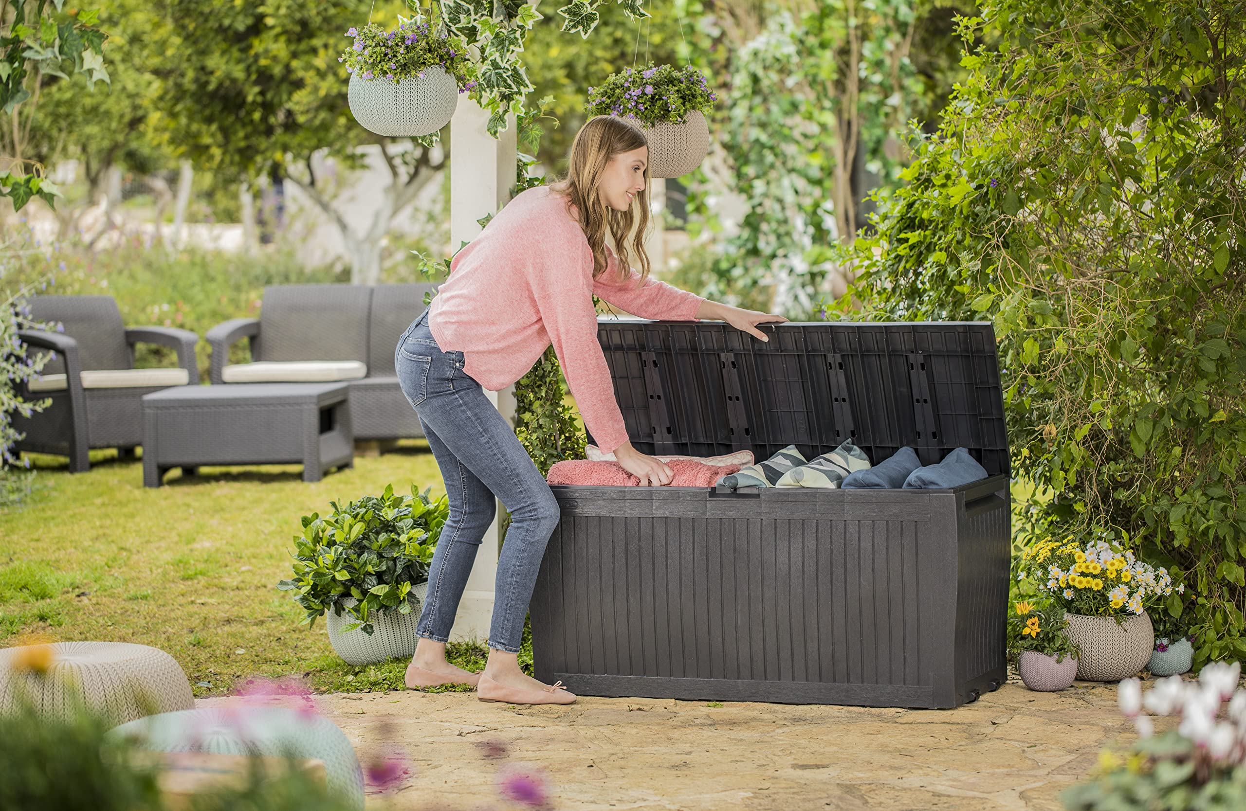 Frau in rosa Pullover öffnet graue Aufbewahrungsbox im Garten mit Pflanzen und Möbeln.