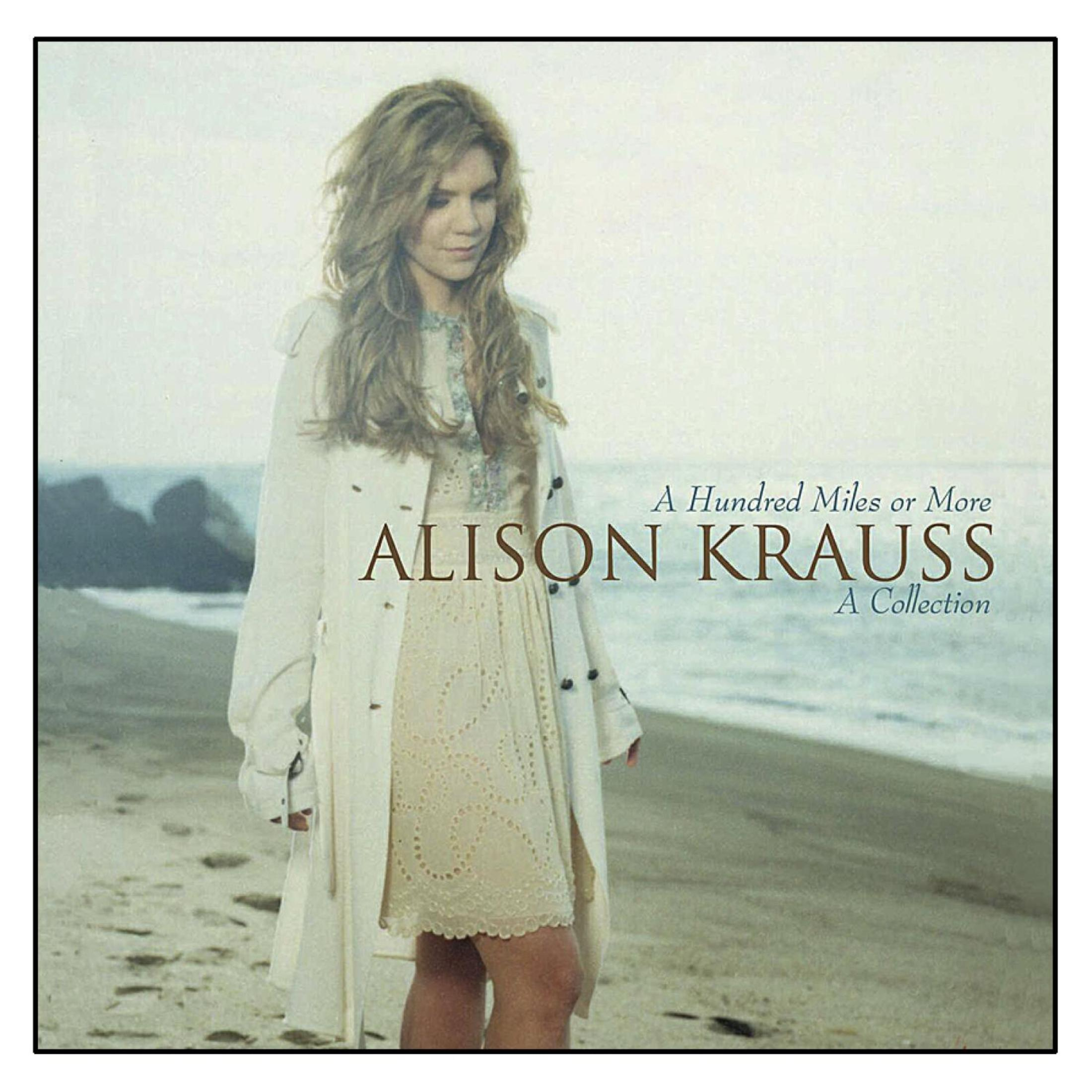 Alison Krauss steht an einem Sandstrand in weißem Mantel und Kleid, im Hintergrund Ozean und Felsen.