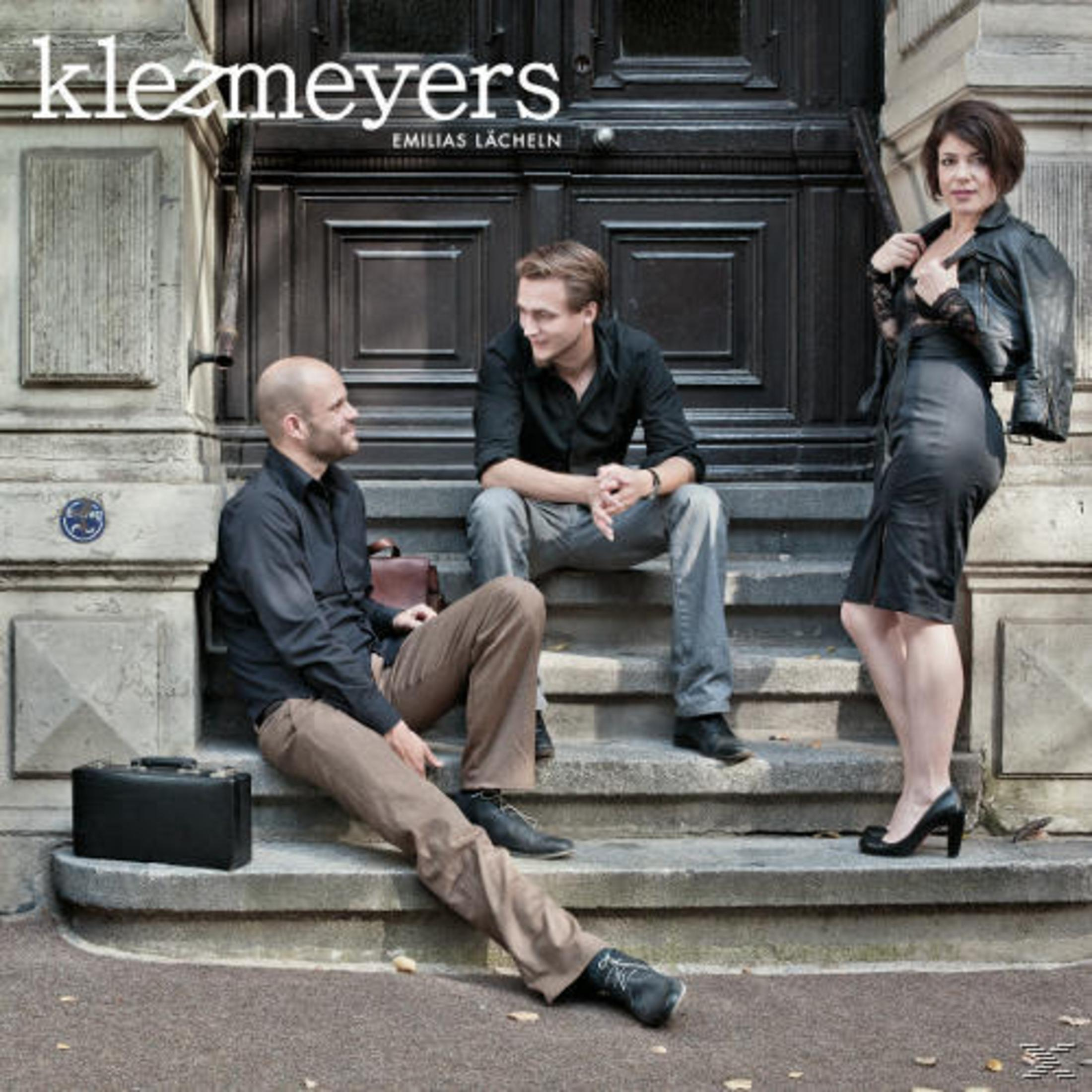 Drei Musiker der Band klezmeyers posieren vor einer schweren Holztür auf Steintreppen.