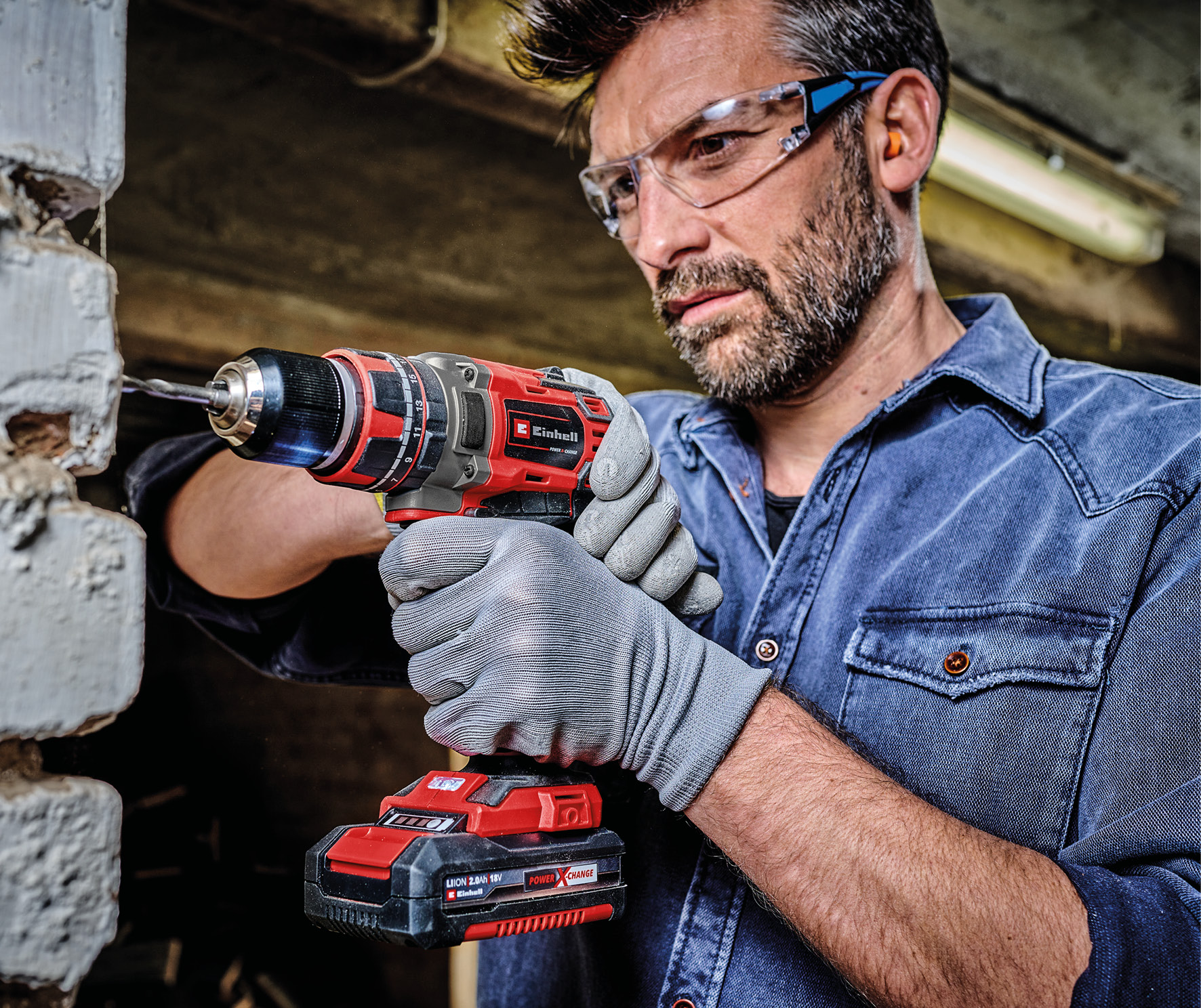 Un lavoratore fora una parete di mattoni con un trapano a batteria rosso, occhiali e guanti.