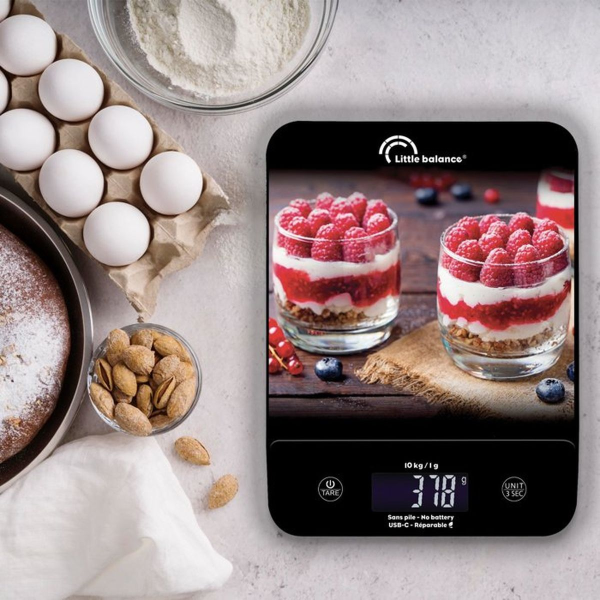 Digitale Küchenwaage mit Himbeerdessert-Bild, umgeben von Eiern, Mandeln, Mehl und einem Kuchen auf der Arbeitsplatte.