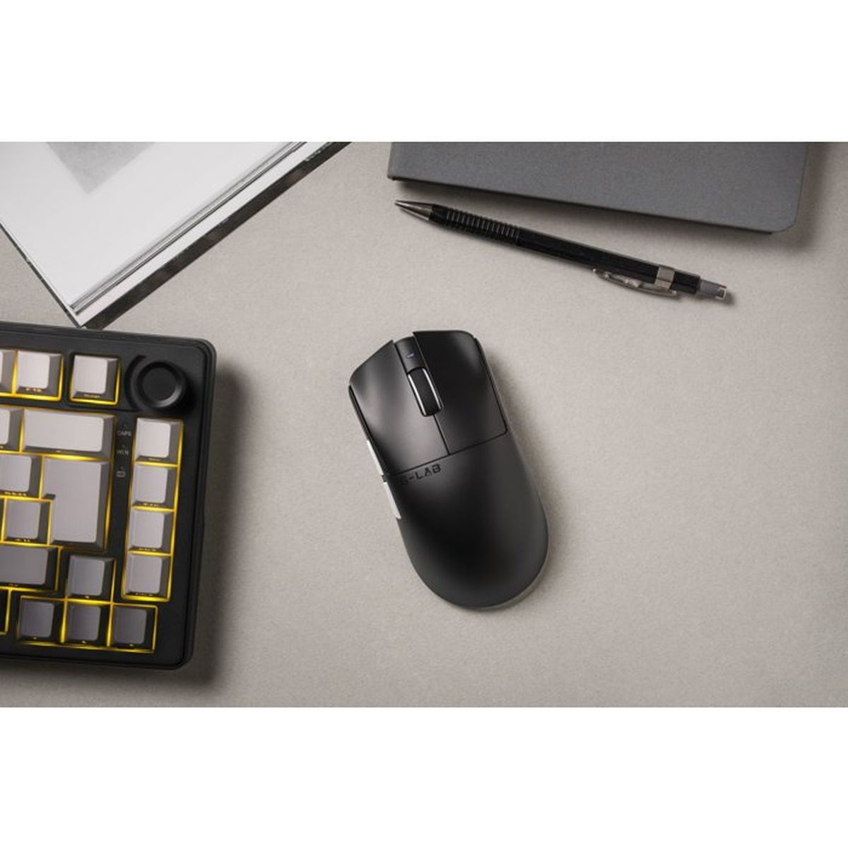 Un ratón inalámbrico negro descansa en un escritorio gris cerca de un teclado iluminado, un bolígrafo y un cuaderno.