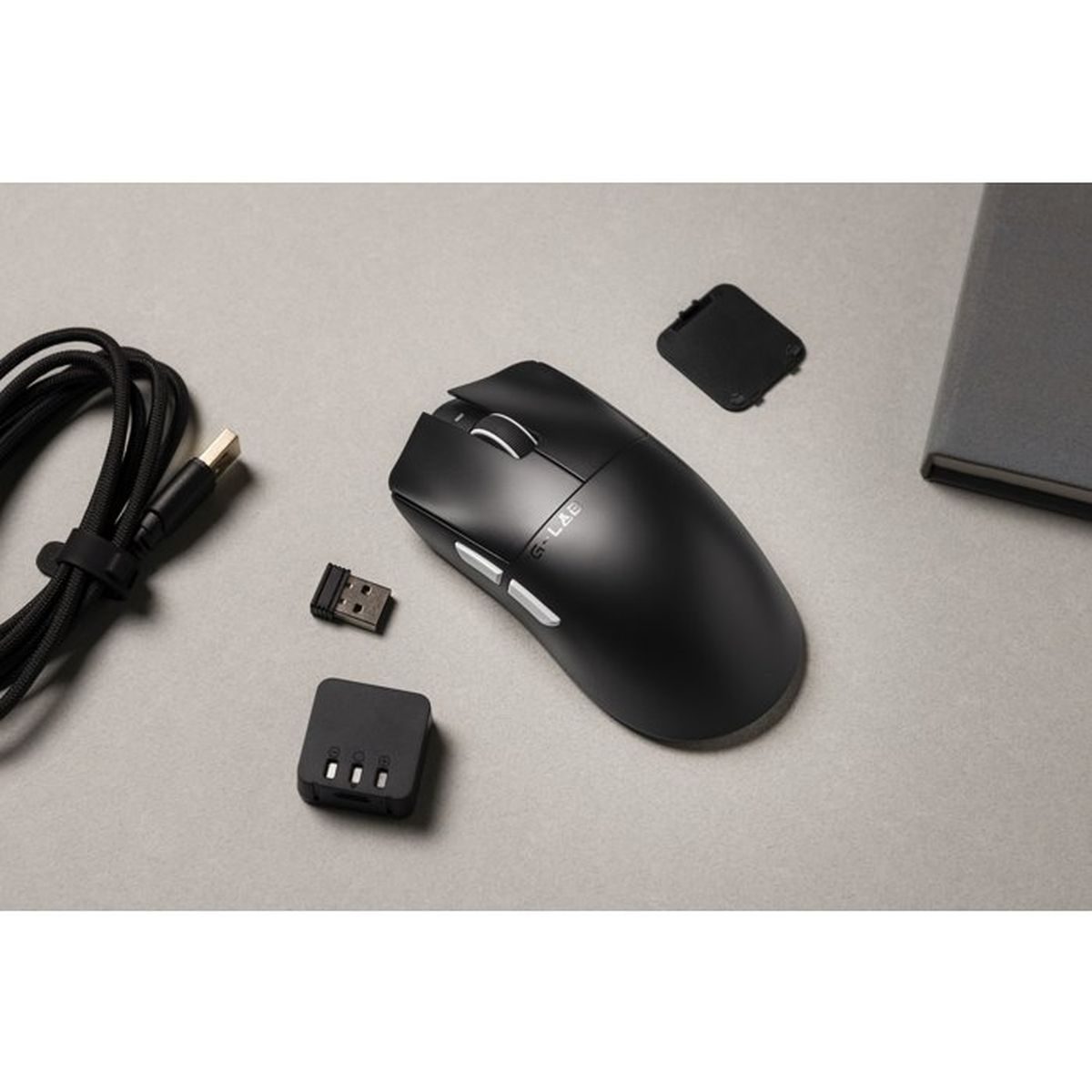 Un ratón inalámbrico negro, un receptor USB, un cable de carga y un cuaderno sobre superficie gris.