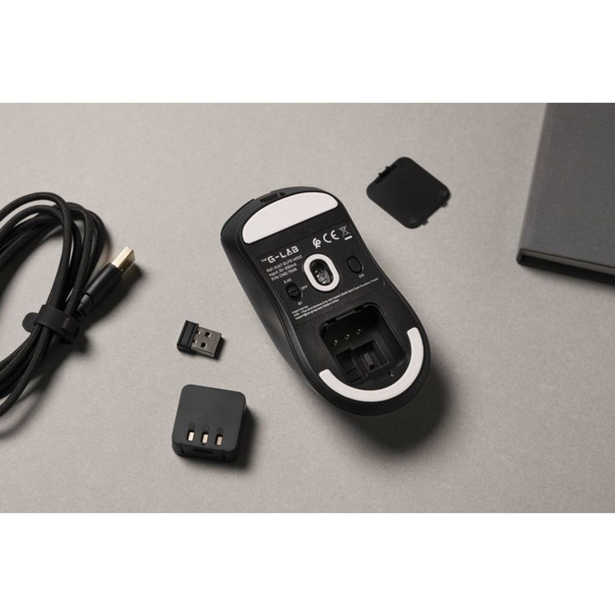 Parte inferior de un ratón de juego inalámbrico negro con accesorios, receptor USB y cable en una mesa gris.
