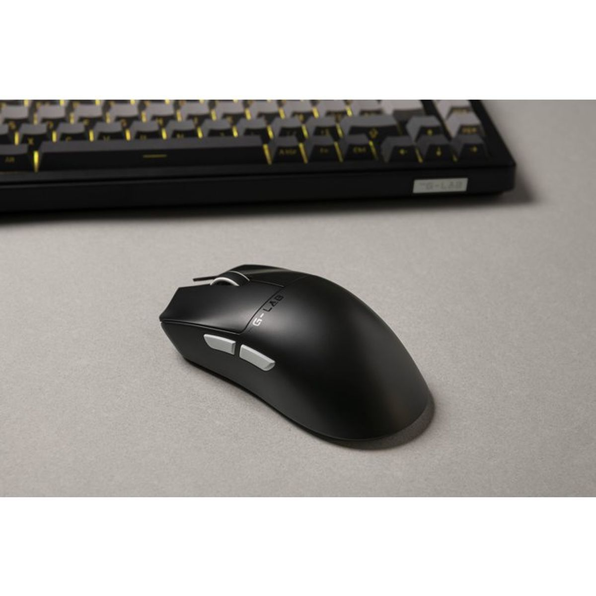 Un ratón de juego negro con botones plateados y logotipo en un escritorio gris, con un teclado al fondo.