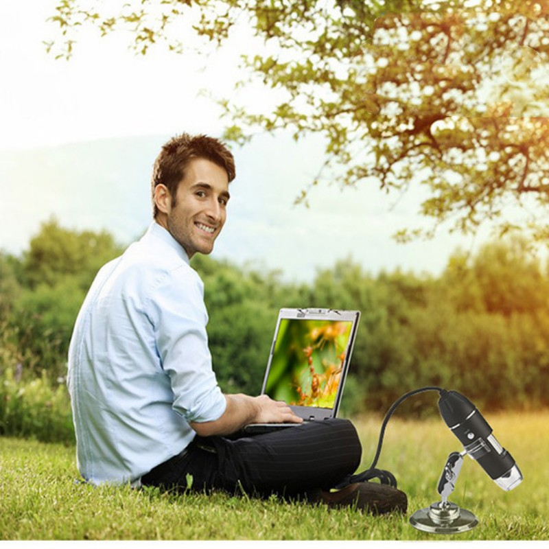Ein Mann sitzt draußen im Gras an einem Laptop, im Vordergrund ein USB-Mikroskop.