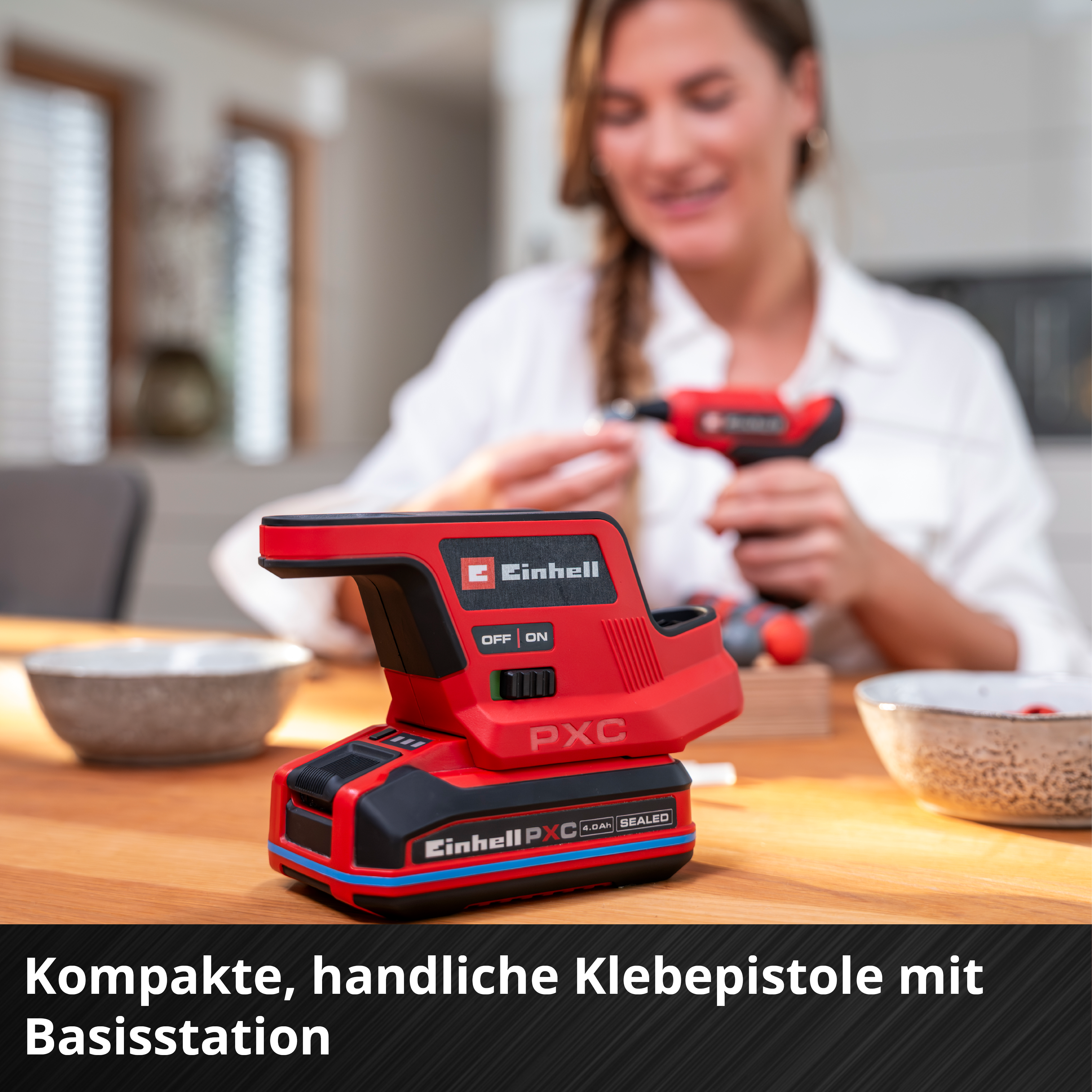 Eine rote Einhell Klebepistole mit Basisstation auf einem Holztisch, im Hintergrund eine Frau.