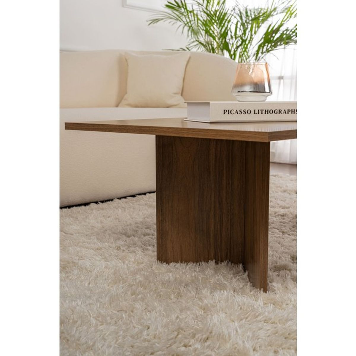 Ein Holzcouchtisch mit Buch und Glas auf einem weißen Hochflorteppich vor einem Beigesofa und einer grünen Pflanze.